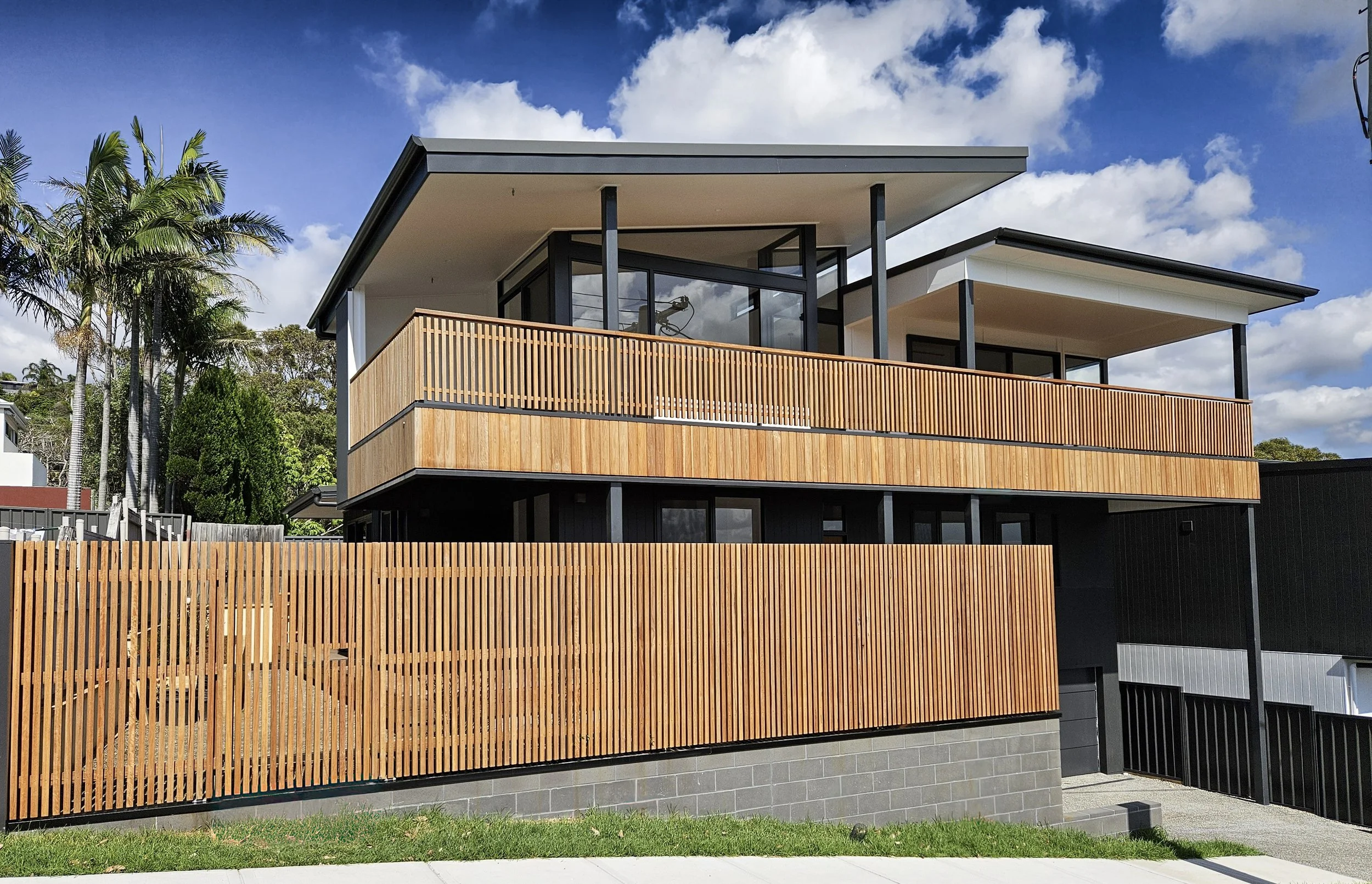Modern two-story house with a black and wood exterior, large windows, and a wooden fence, set against a partly cloudy sky.