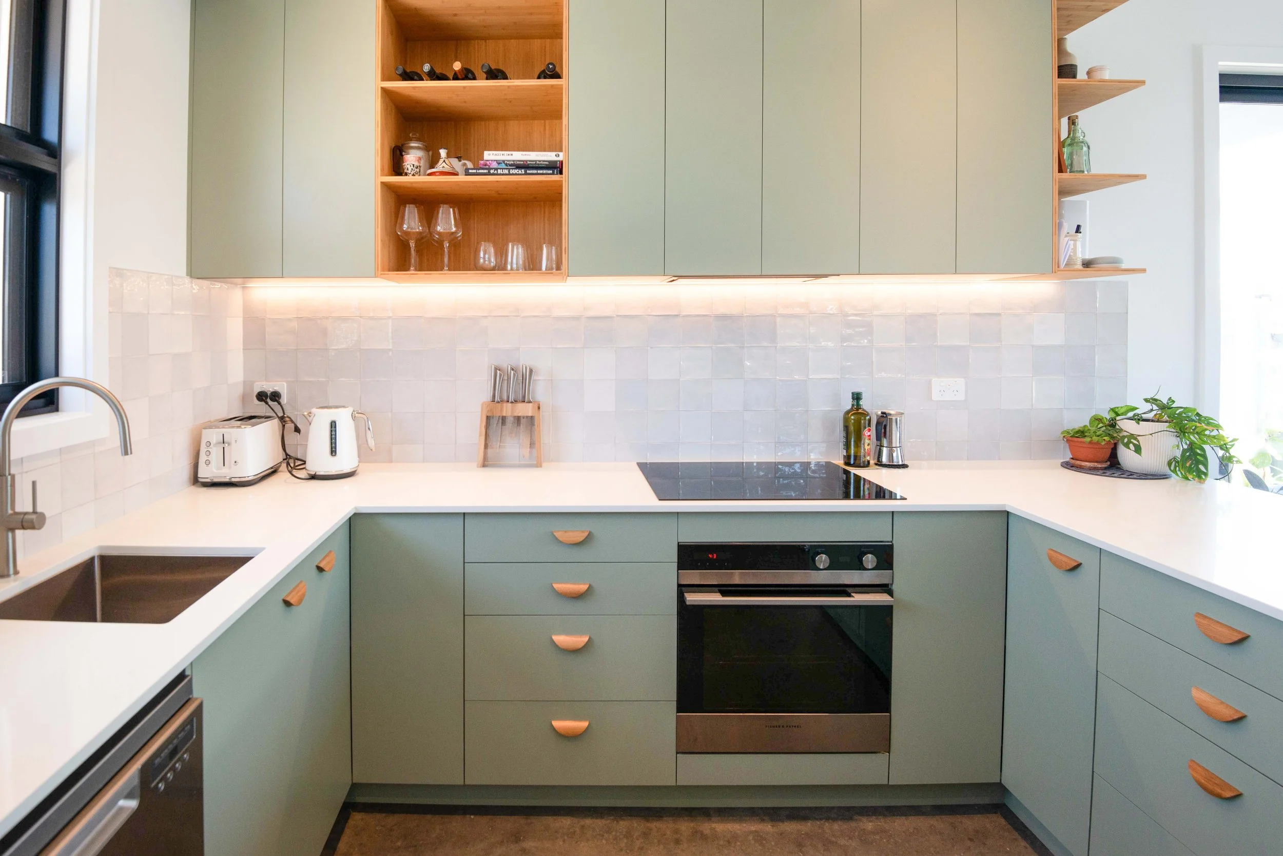 Modern kitchen with green cabinets, white countertops, and a white tile backsplash. Kitchen appliances include a toaster, electric kettle, and oven. There are open shelves with glasses, plants, and decorative items.