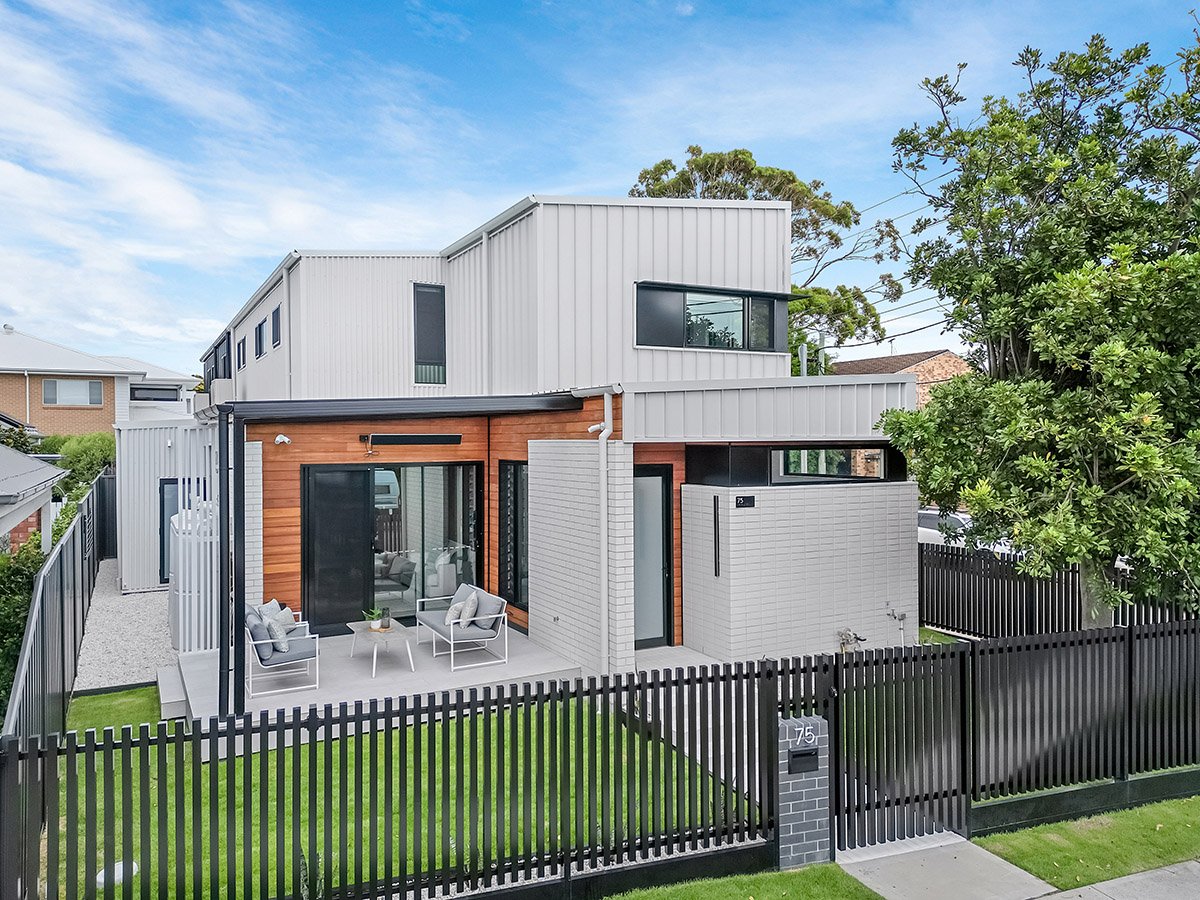 Modern two-story house with a small front yard, black fence, and outdoor seating area, featuring a mix of wooden and metal siding, surrounded by trees and neighboring houses.