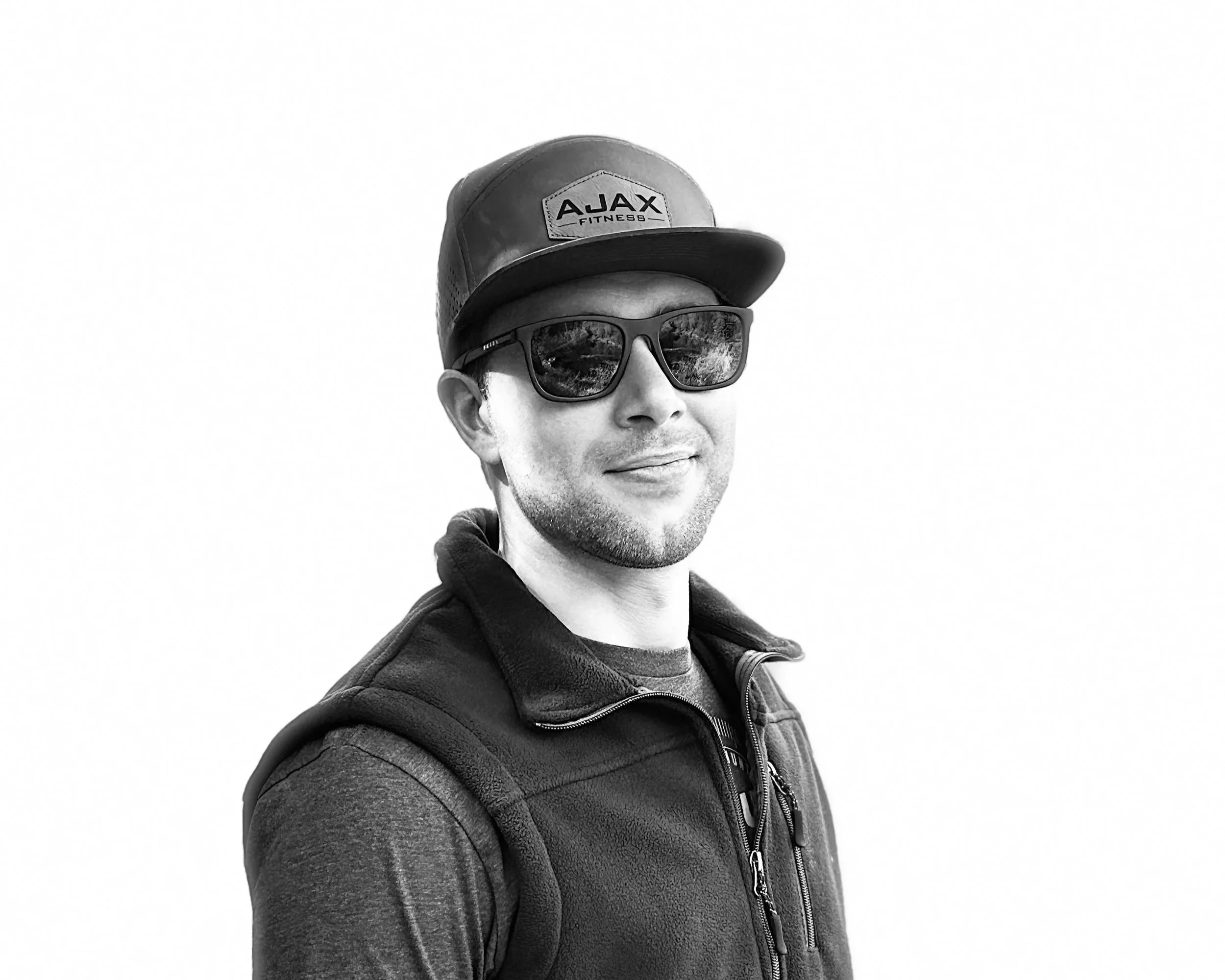 A man wearing a baseball cap with 'AJAX' written on it, sunglasses, a zip-up jacket, and a backpack, smiling outdoors against a plain background.