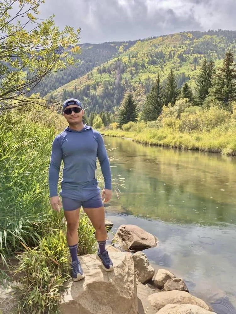 A person in outdoor athletic clothing stands on rocks beside a river, surrounded by green trees and mountains, under a cloudy sky.