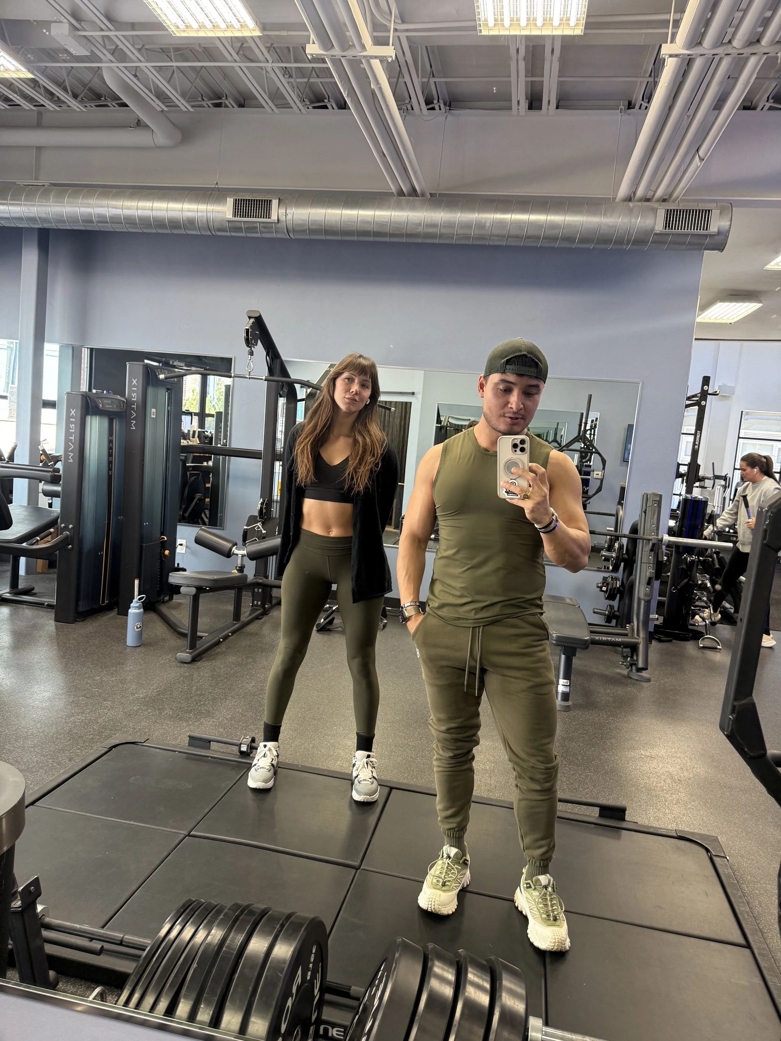 A man and a woman taking a mirror selfie in a gym. The man is wearing a green sleeveless shirt, green joggers, and a backwards cap, while the woman is wearing a black sports bra, green leggings, and a black jacket.