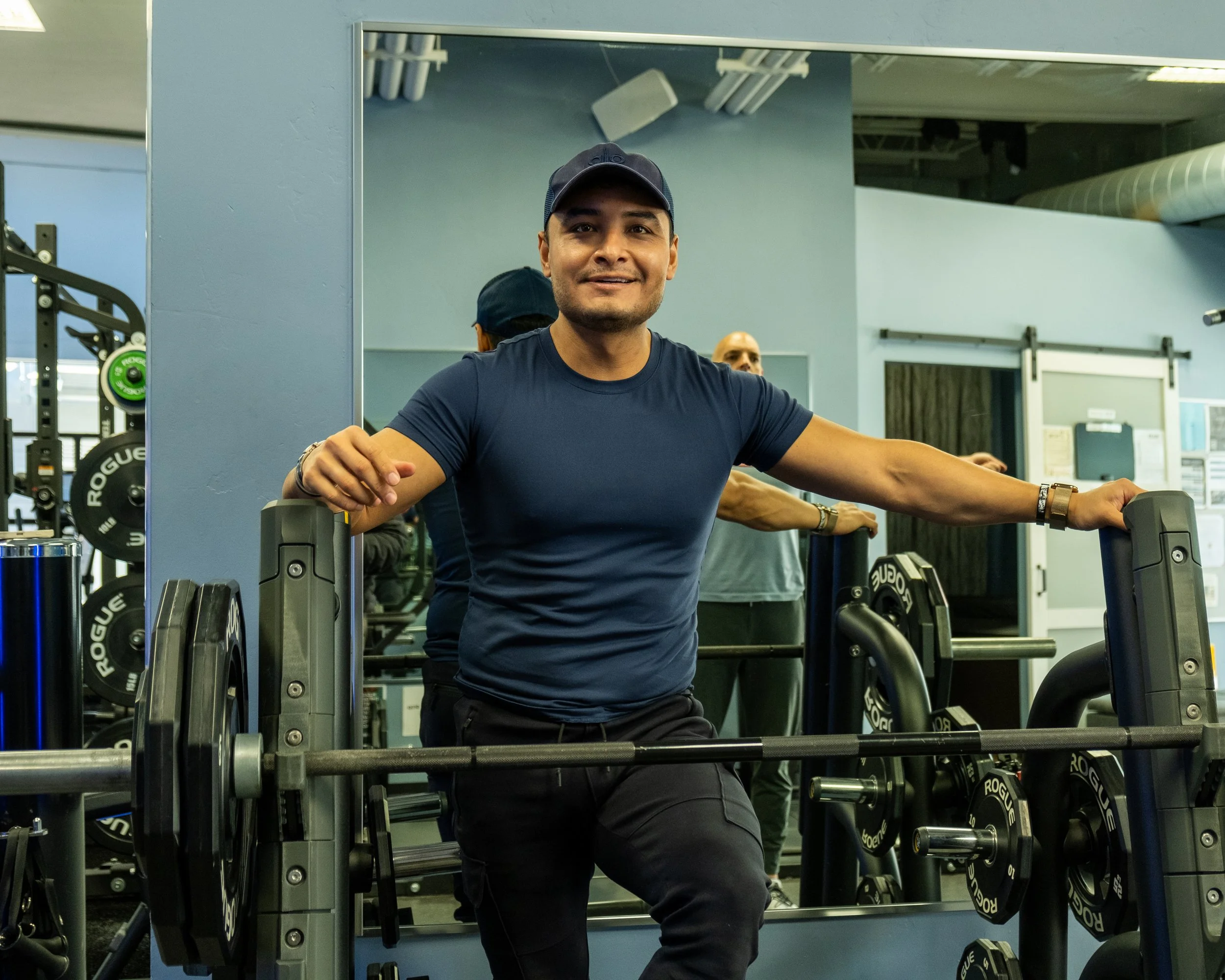 A man in a blue shirt and cap standing at a gym station with weight plates, smiling at the camera.