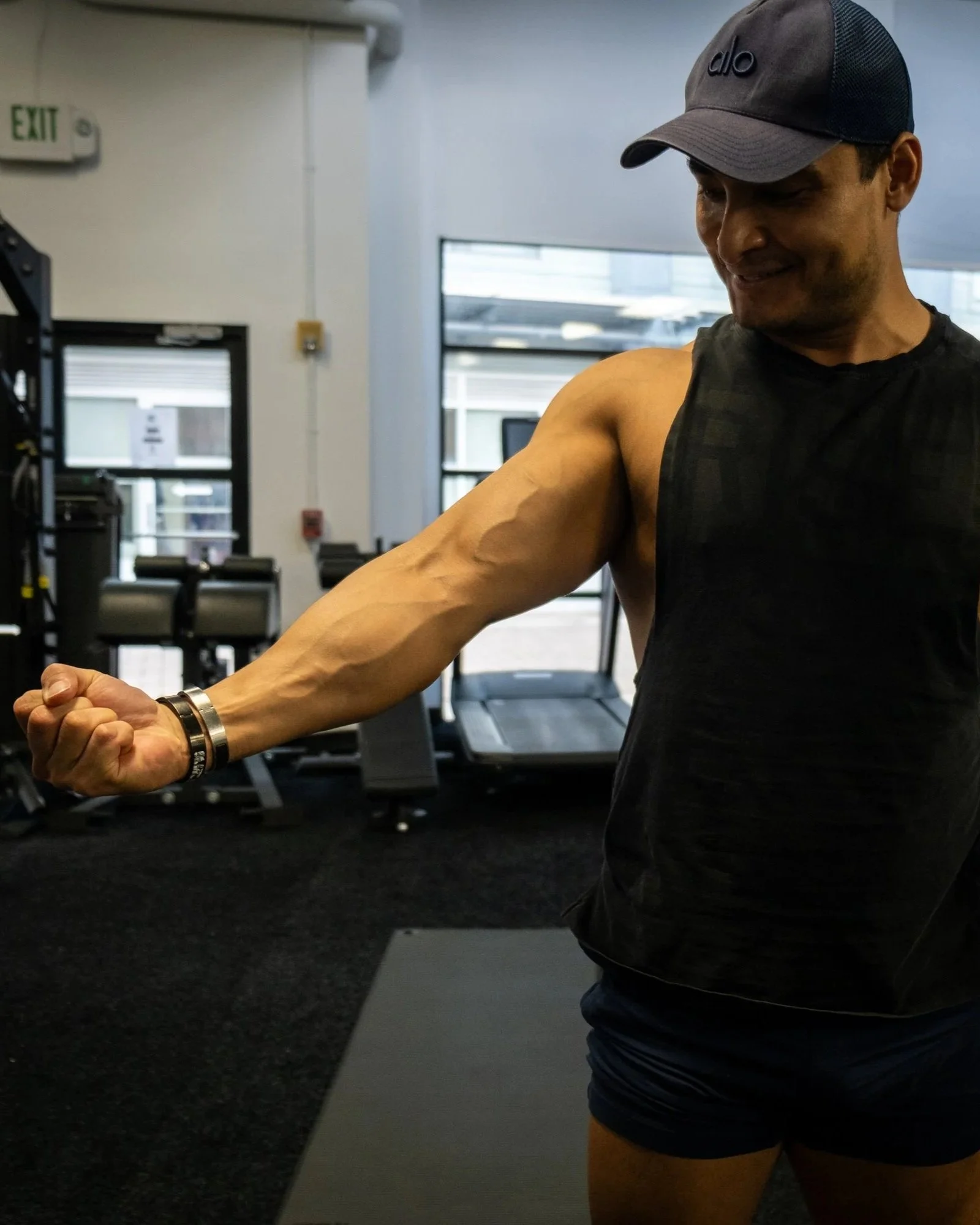 A man in a black sleeveless shirt and black shorts, wearing a gray cap, is flexing his arm in a gym with weightlifting equipment in the background.