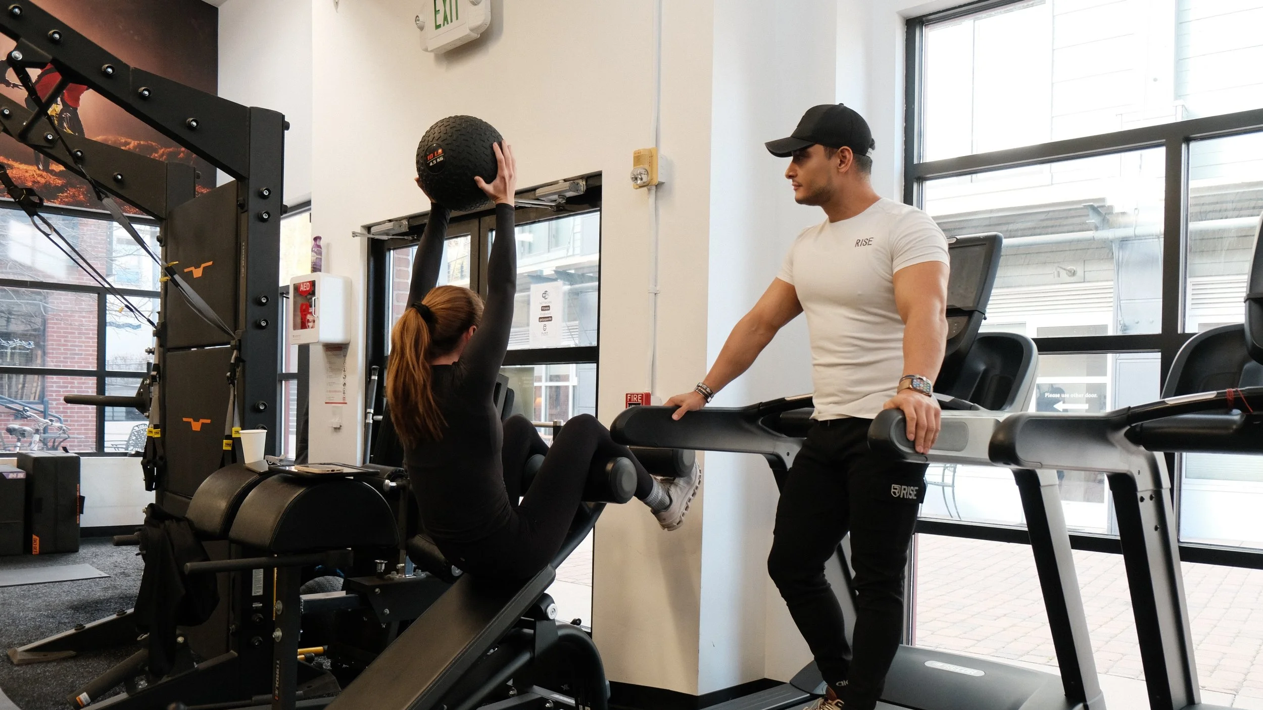 A woman exercises on a rowing machine in a gym, supervised by a trainer on a treadmill, with large windows in the background.