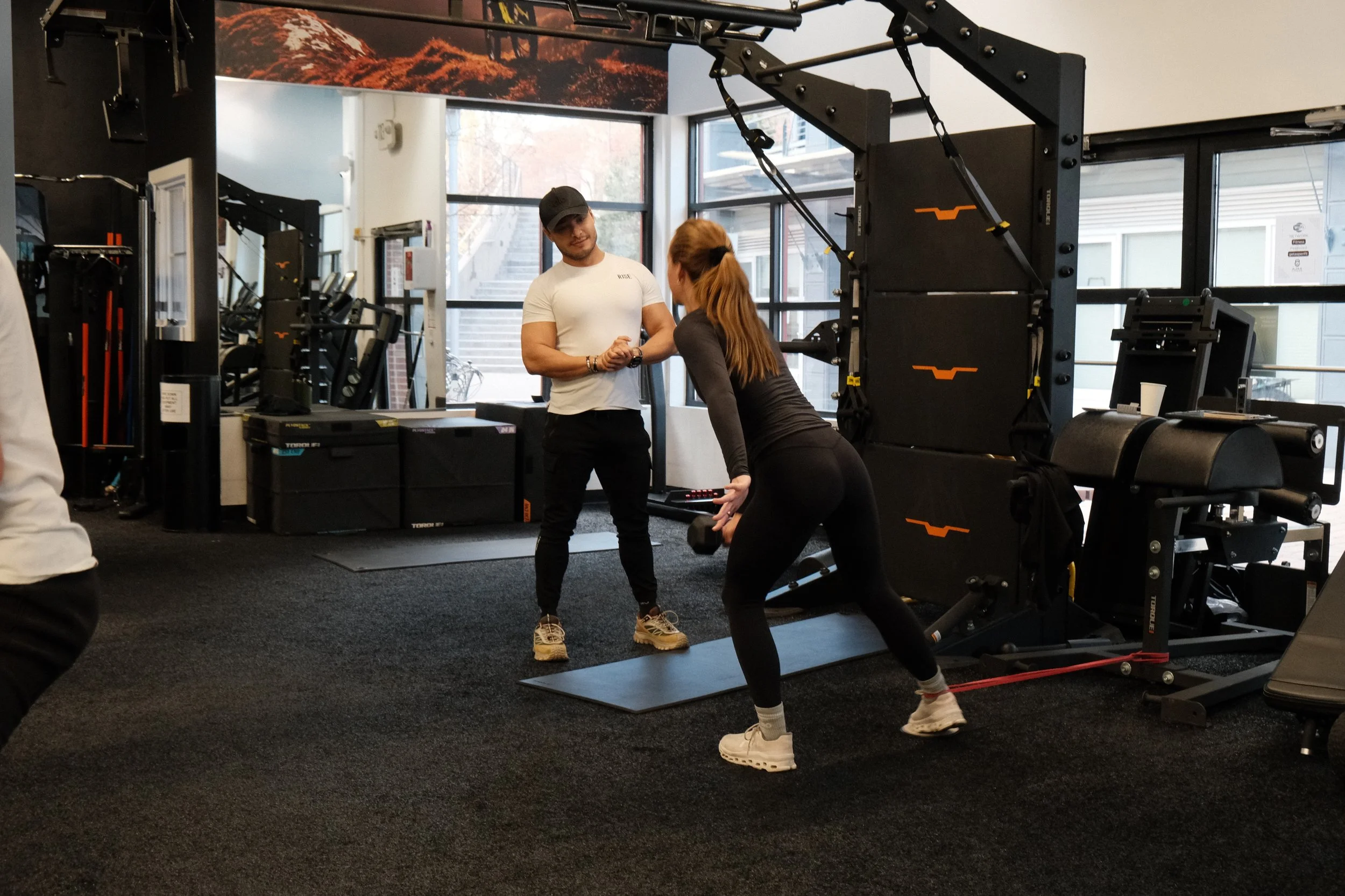 A woman exercises with a dumbbell while a trainer assists, in a gym with fitness equipment and large windows.