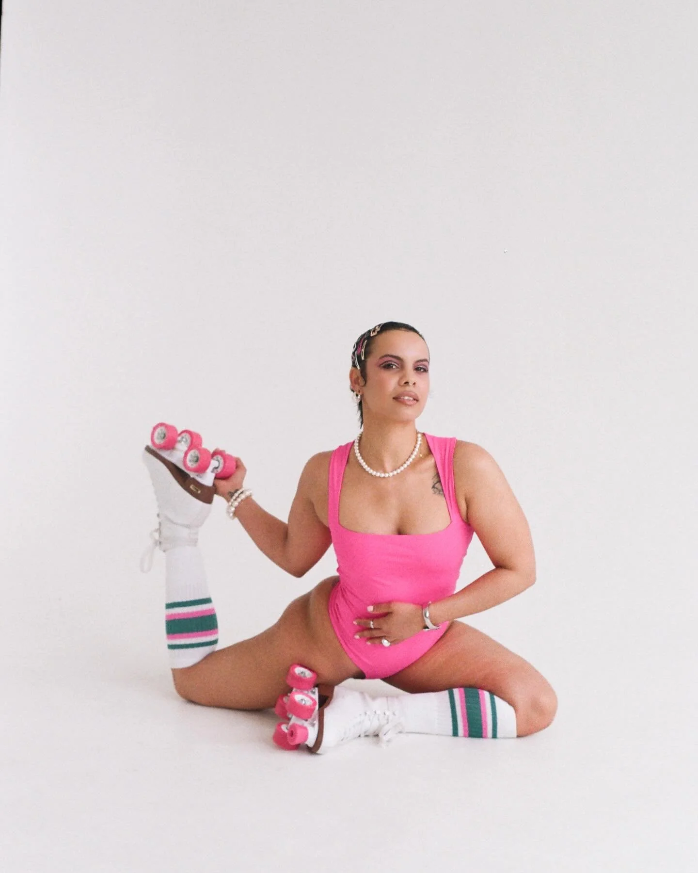 A woman dressed in pink with striped socks poses with one leg raised and holding pink roller skates, against a plain white background.