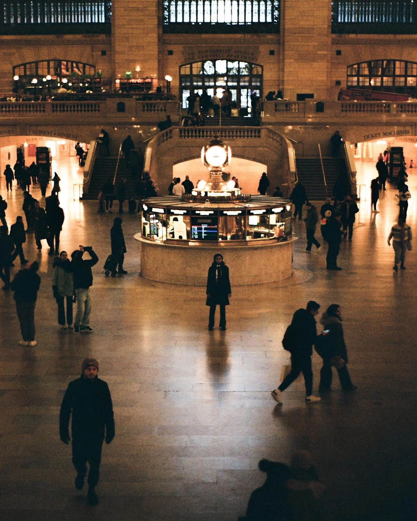 Interior of a large train station with people walking and standing, a central information desk, escalators, and an upper level with more people visible through large windows.