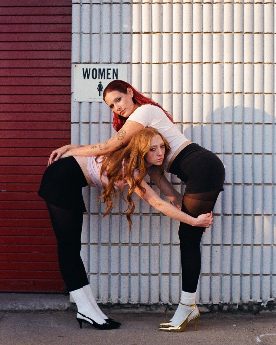 Two women with red hair and tattoos, dressed in black and white, leaning against a wall with a 'Women' restroom sign in the background.