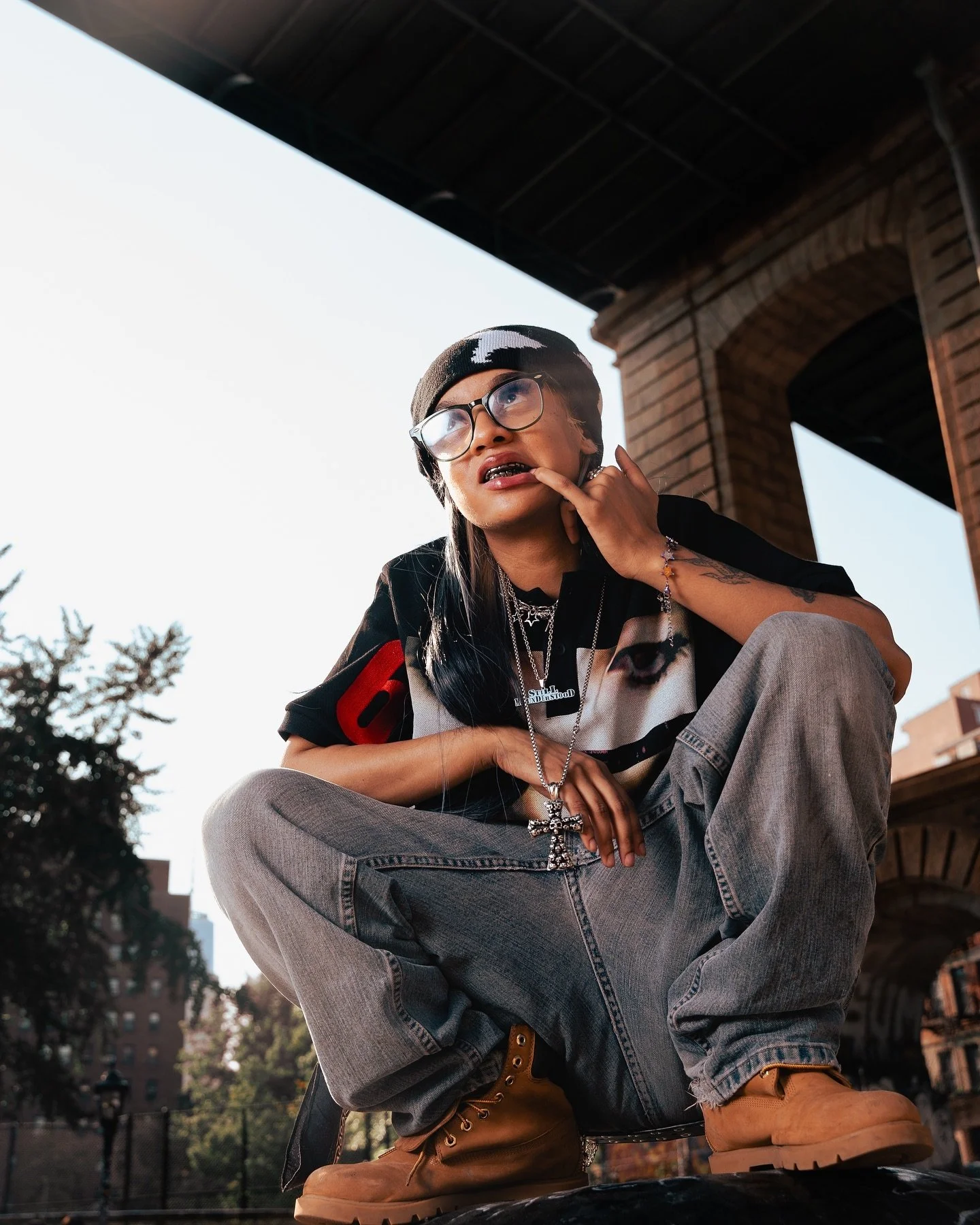 Young adult wearing glasses, beanie, and layered jewelry, squatting outdoors near a brick building, with trees and urban environment in the background.