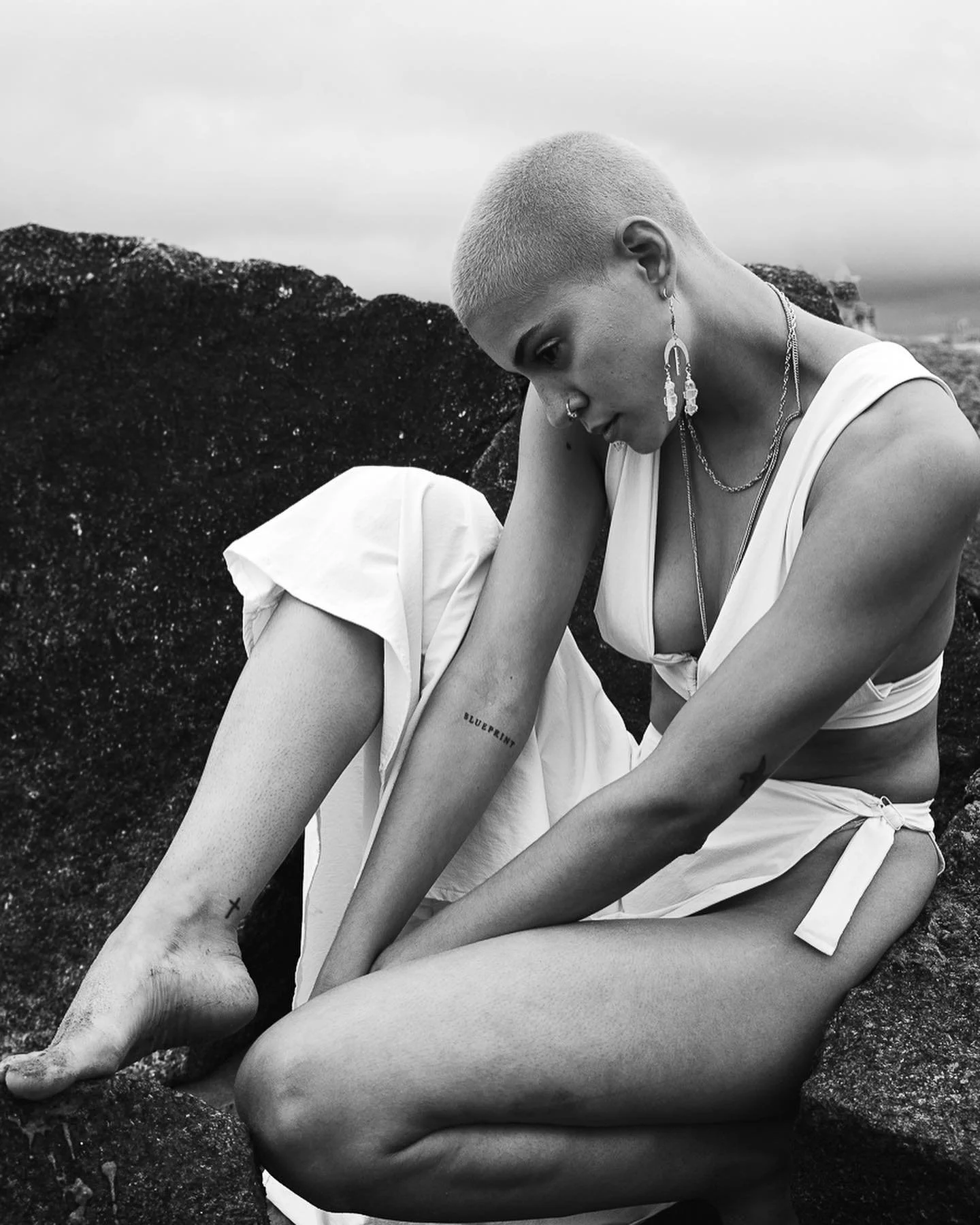 A black and white photo of a woman with blond hair sitting on rocks at the beach. She wears a sleeveless top, shorts, and multiple necklaces with long earrings. She has a tattoo that reads 'BLUEPRINT' on her thigh and a small tattoo of a cross on her ankle. The background shows cloudy skies and part of the sea.