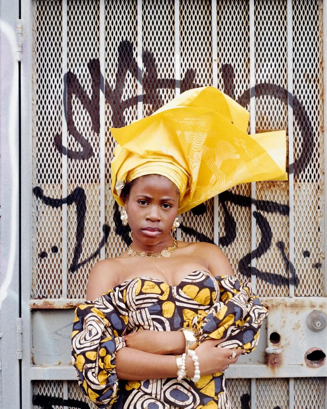 A woman standing with arms crossed in front of a metal gate with graffiti, wearing traditional African clothing and a large yellow headwrap.