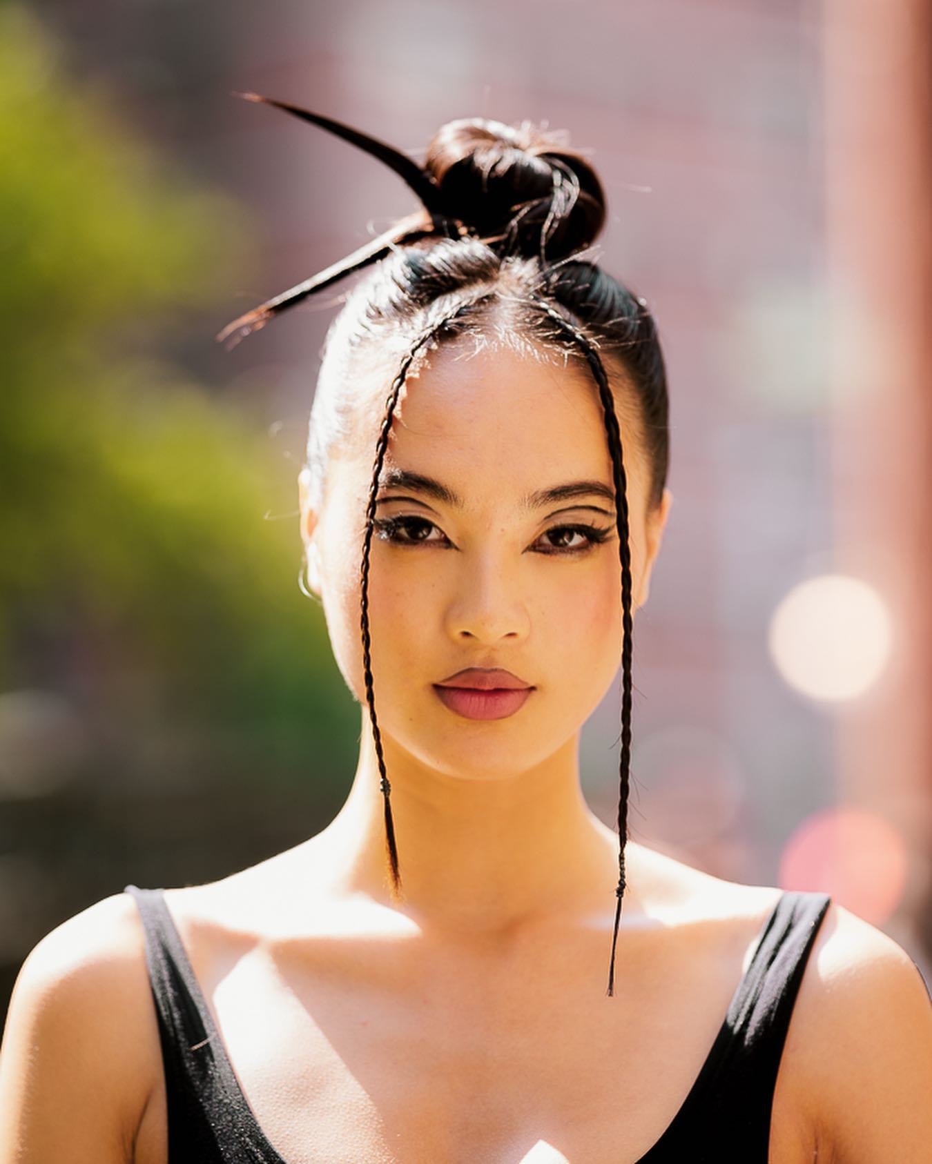 Close-up of a woman with dark hair styled in a high bun with long braided strands, wearing a black spaghetti strap top, outdoors in natural sunlight.