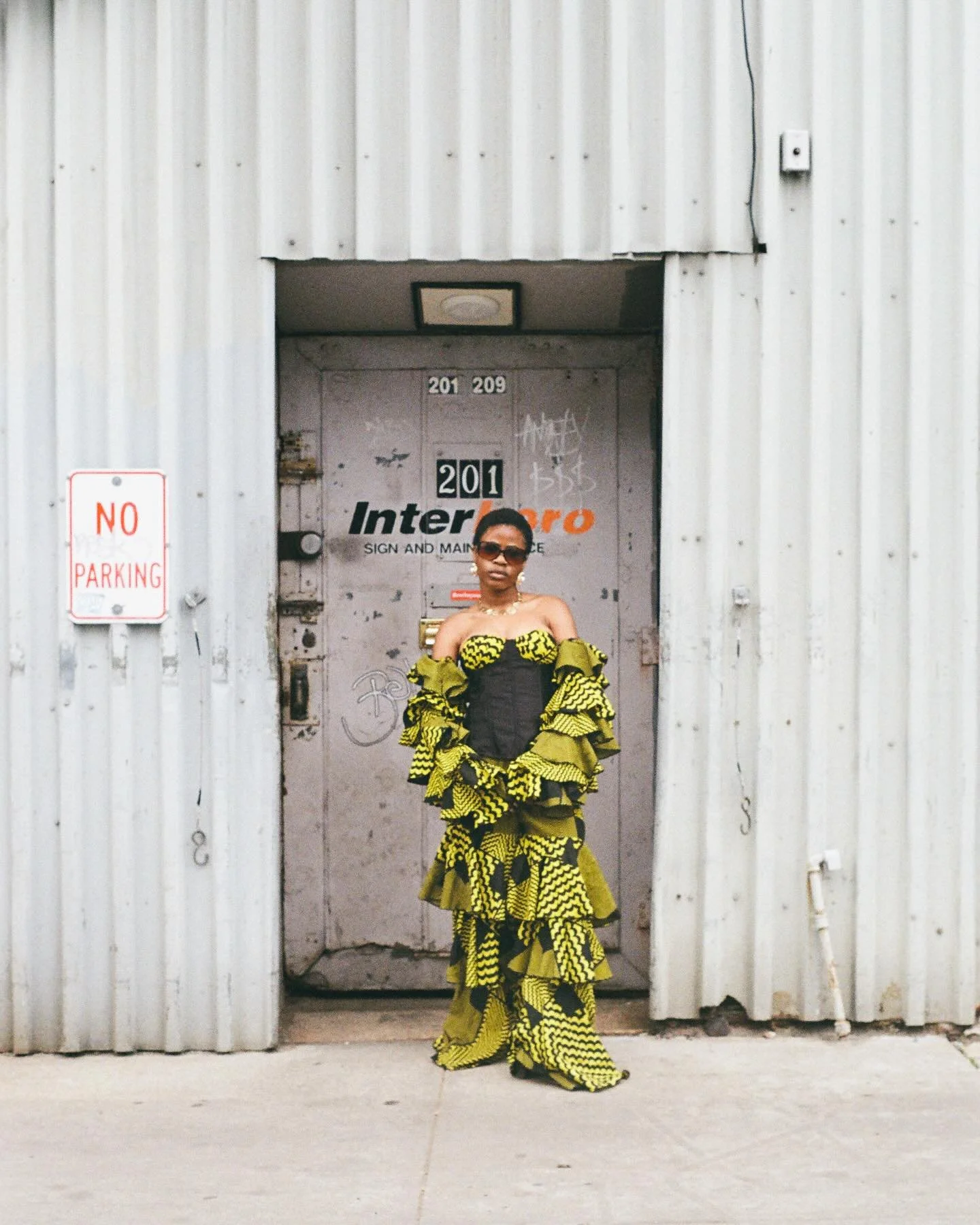 A woman wearing sunglasses in front of a gray industrial door with yellow and black patterned ruffled dress.