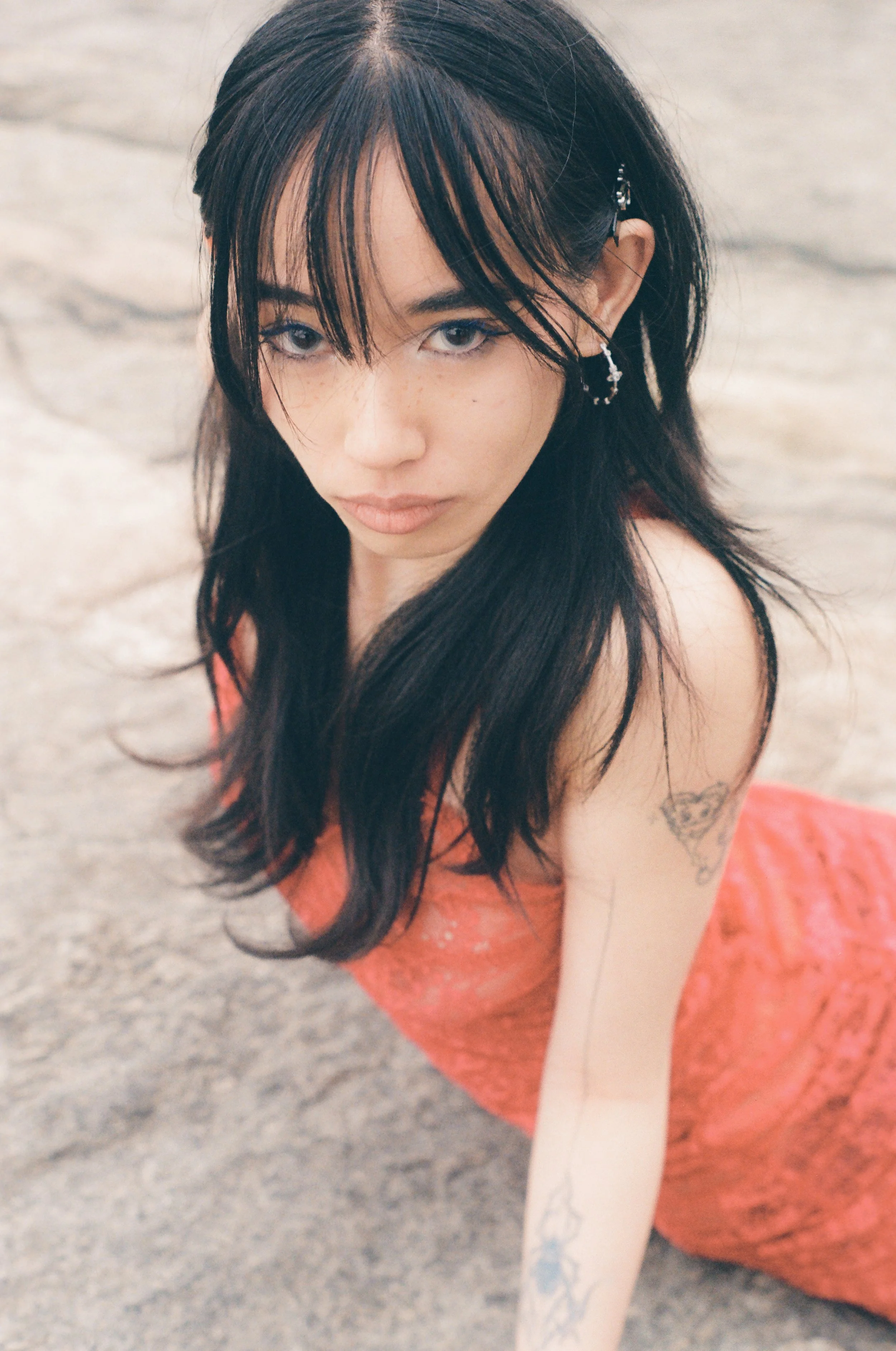 A woman with long dark hair and a nose piercing, wearing a coral-colored dress, sitting on a beach with sandy ground visible.