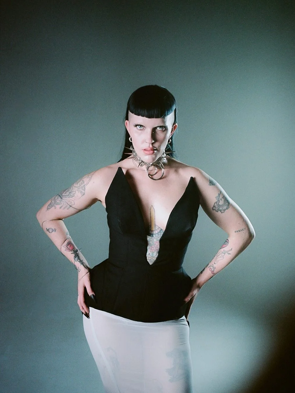 Young woman with black hair, tattoos, and dark makeup poses with hands on hips against a plain background.