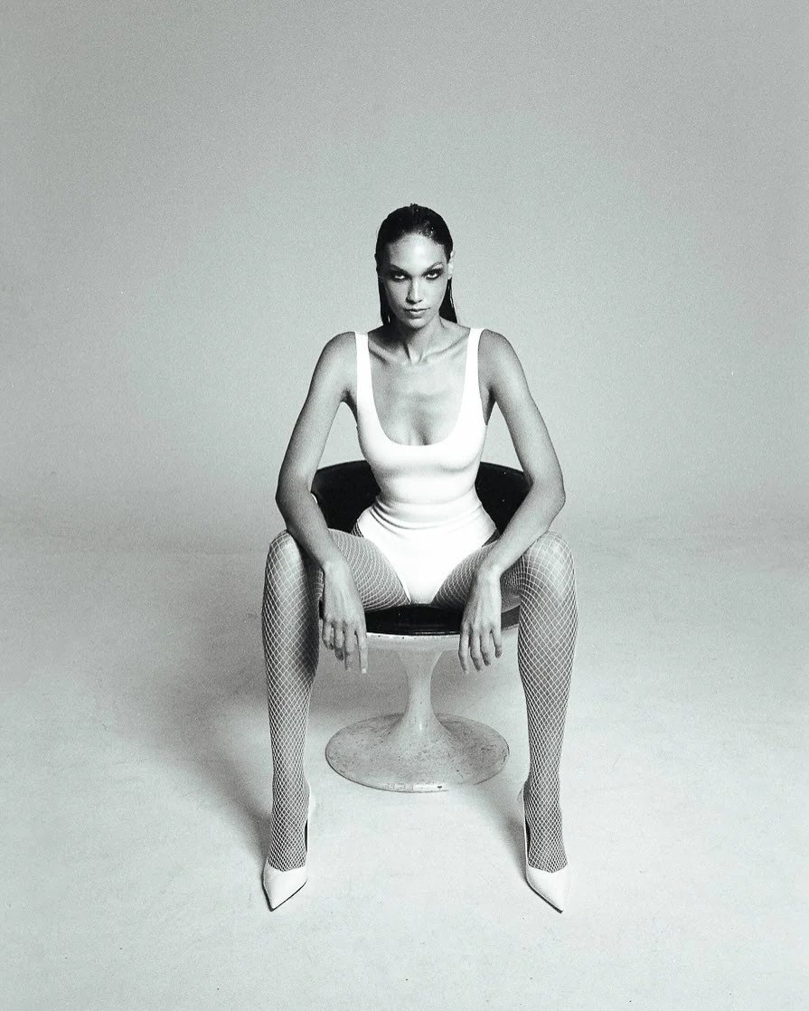A woman in fishnet stockings and a fitted sleeveless dress sitting on a modern chair in a minimalist setting.