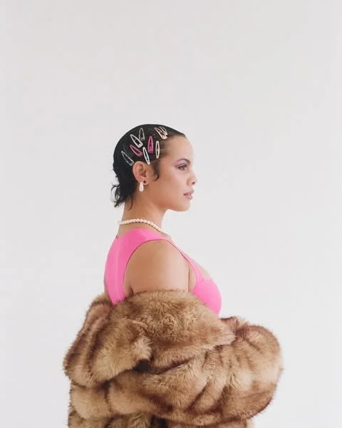Side profile of a woman with short dark hair decorated with colorful hair clips, wearing a pearl necklace, pink top, and a fur coat, against a plain white background.