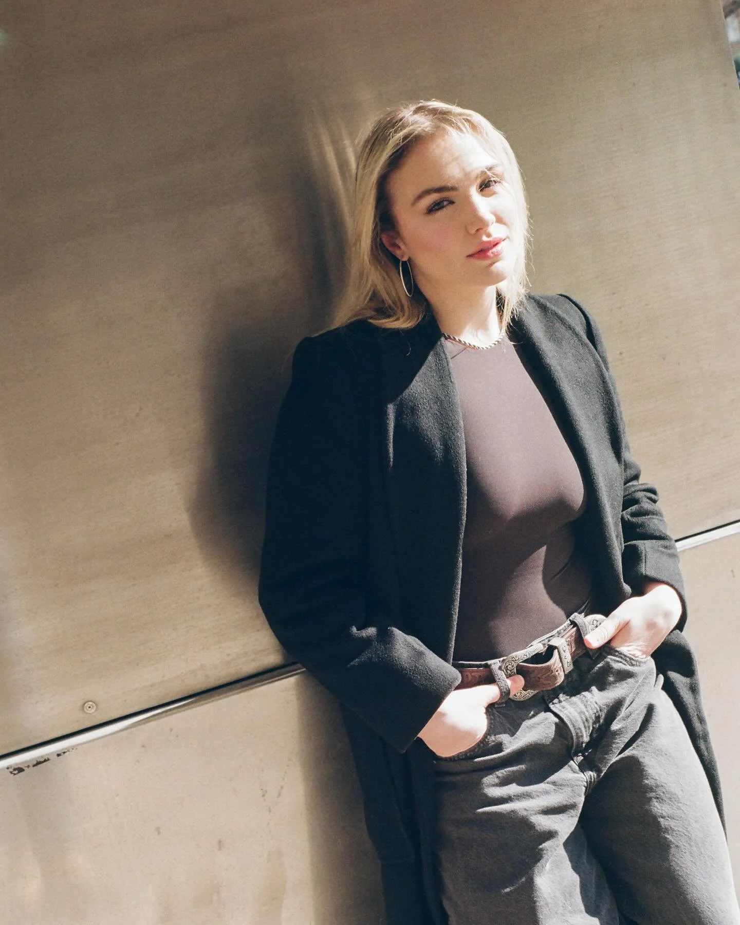 A young woman with blonde hair leaning against a metallic wall, wearing a dark blazer, black sheer top, and loose gray pants, holding her belt with one hand and looking at the camera.