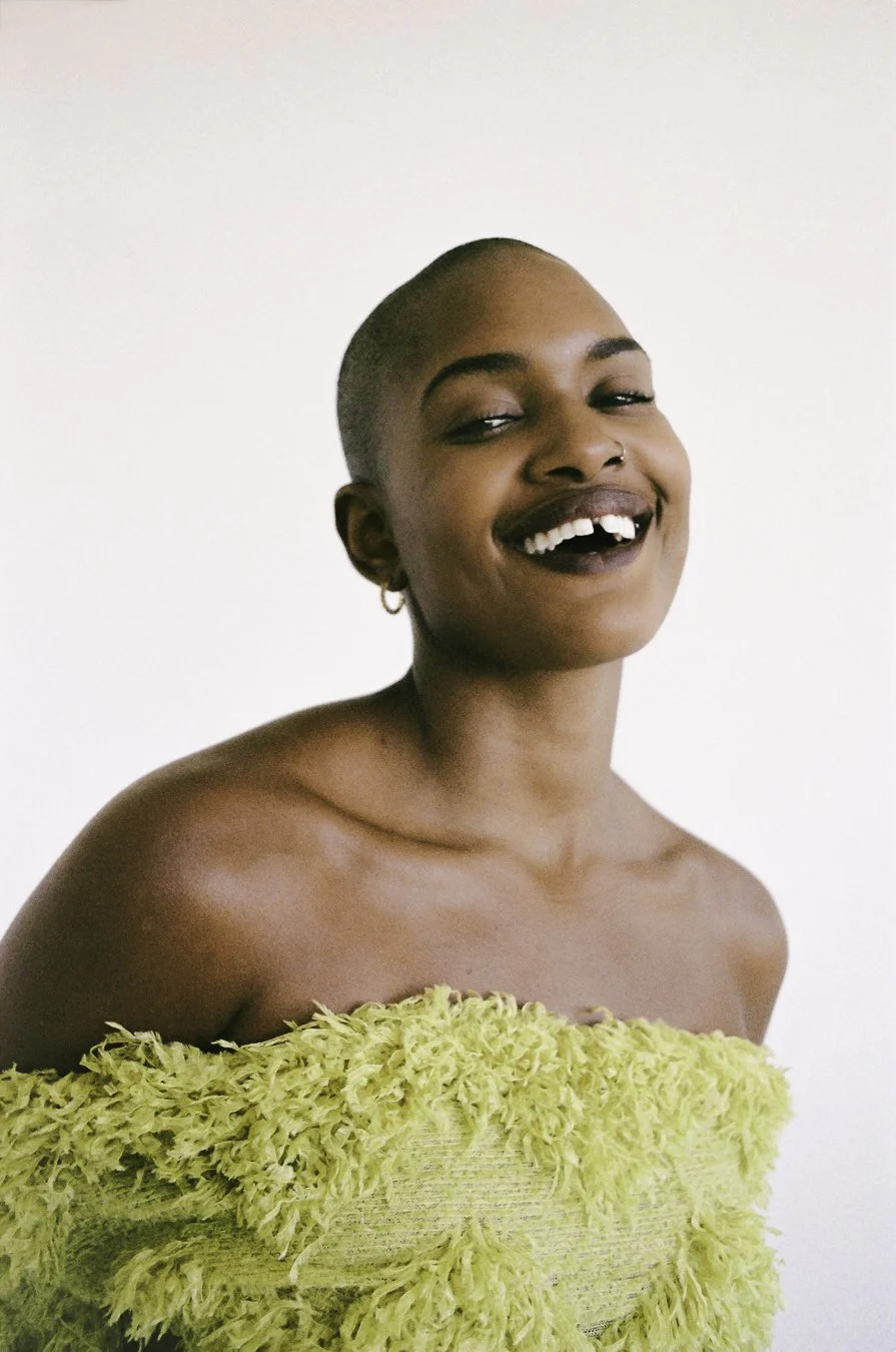 A young person with a shaved head smiling and showing missing teeth, wearing a yellow textured off-the-shoulder top against a plain white background.