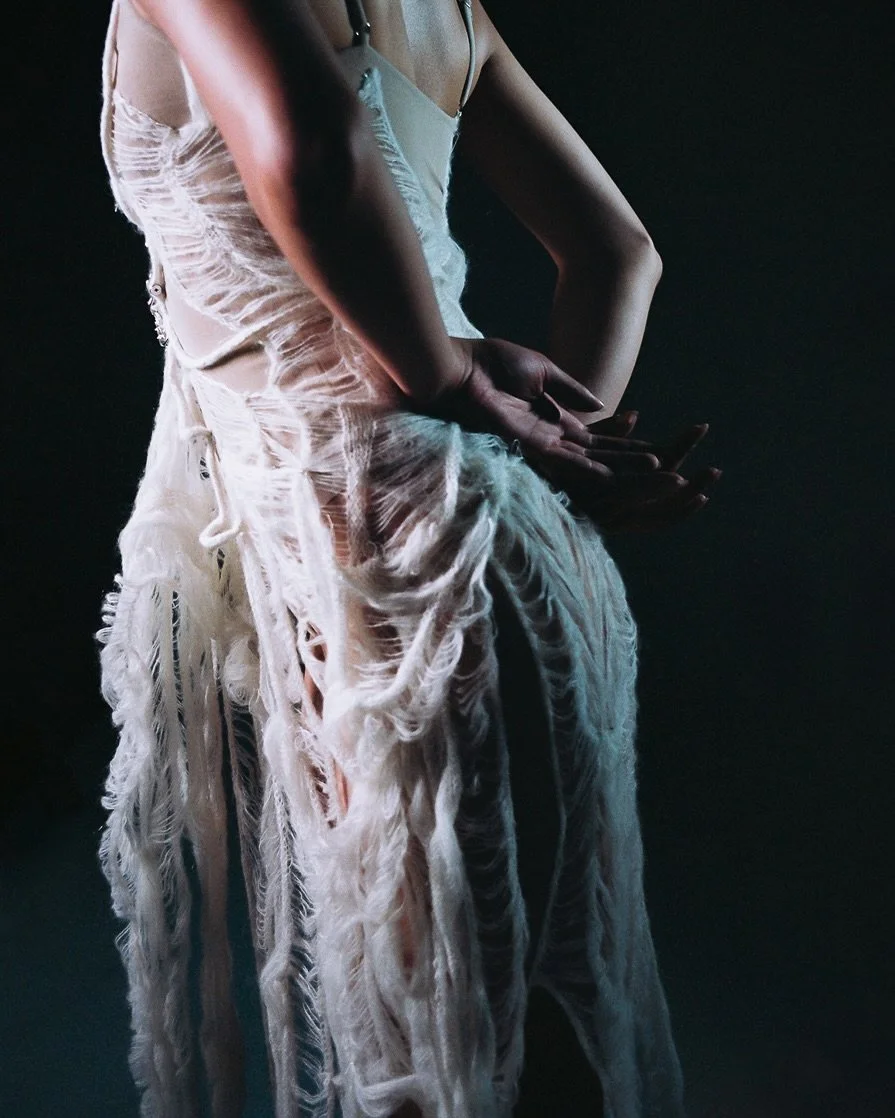 Close-up of a person wearing a distressed, torn white dress, with hands resting on their waist against a dark background.
