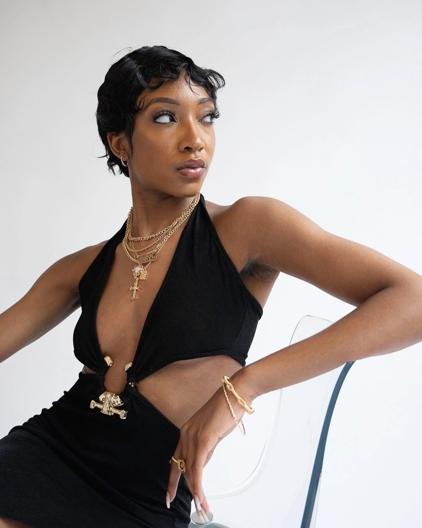 A woman with short black hair wearing a black sleeveless top with gold jewelry, including necklaces and bracelets, sitting on a chair against a plain white background.
