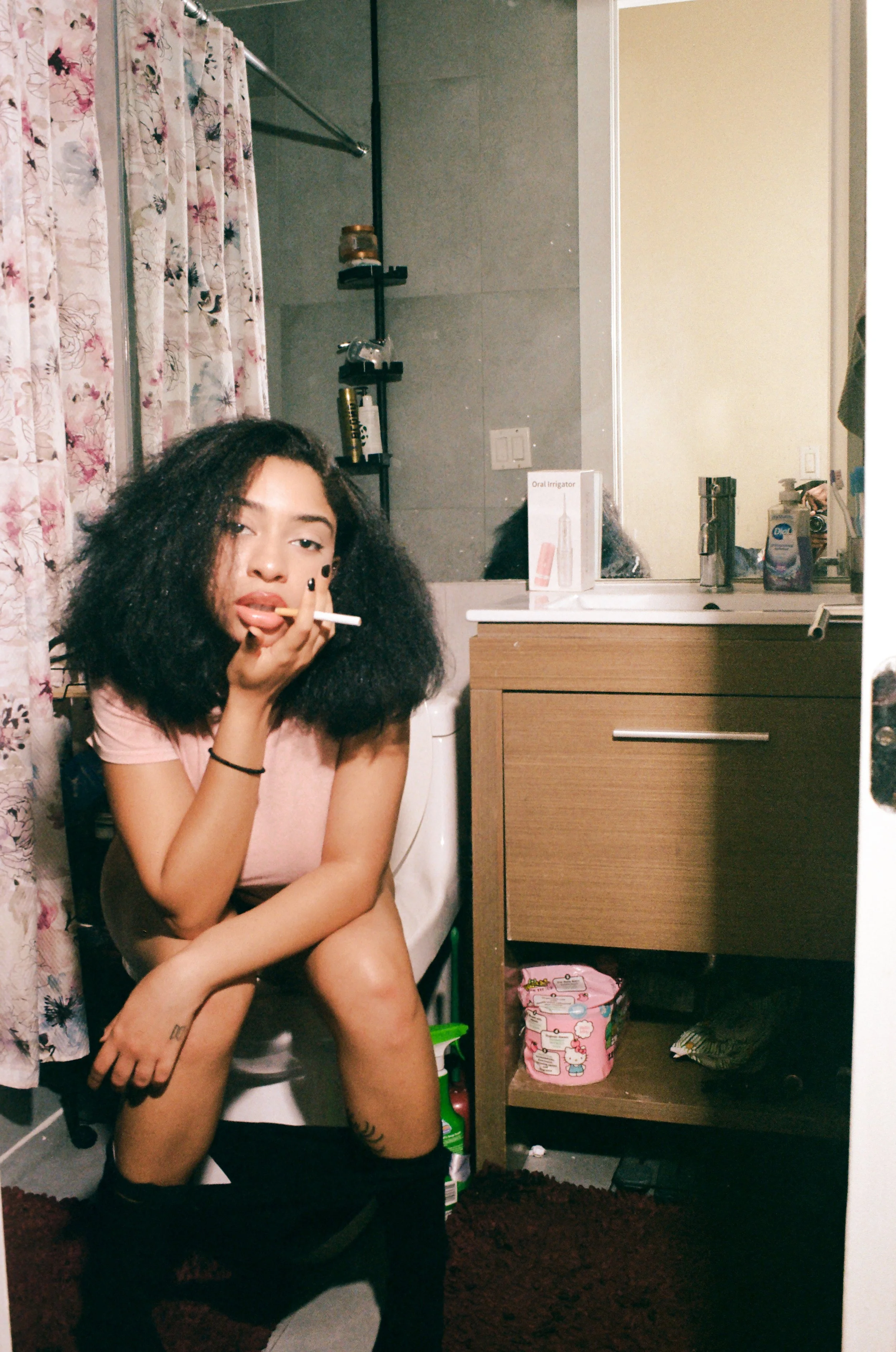 A young woman with curly hair sitting on a toilet in a bathroom, holding a cigarette to her lips, with her arm resting on another person's shoulder. The bathroom has floral curtains, a dark towel, and various toiletries on a shelf and on the sink. Th