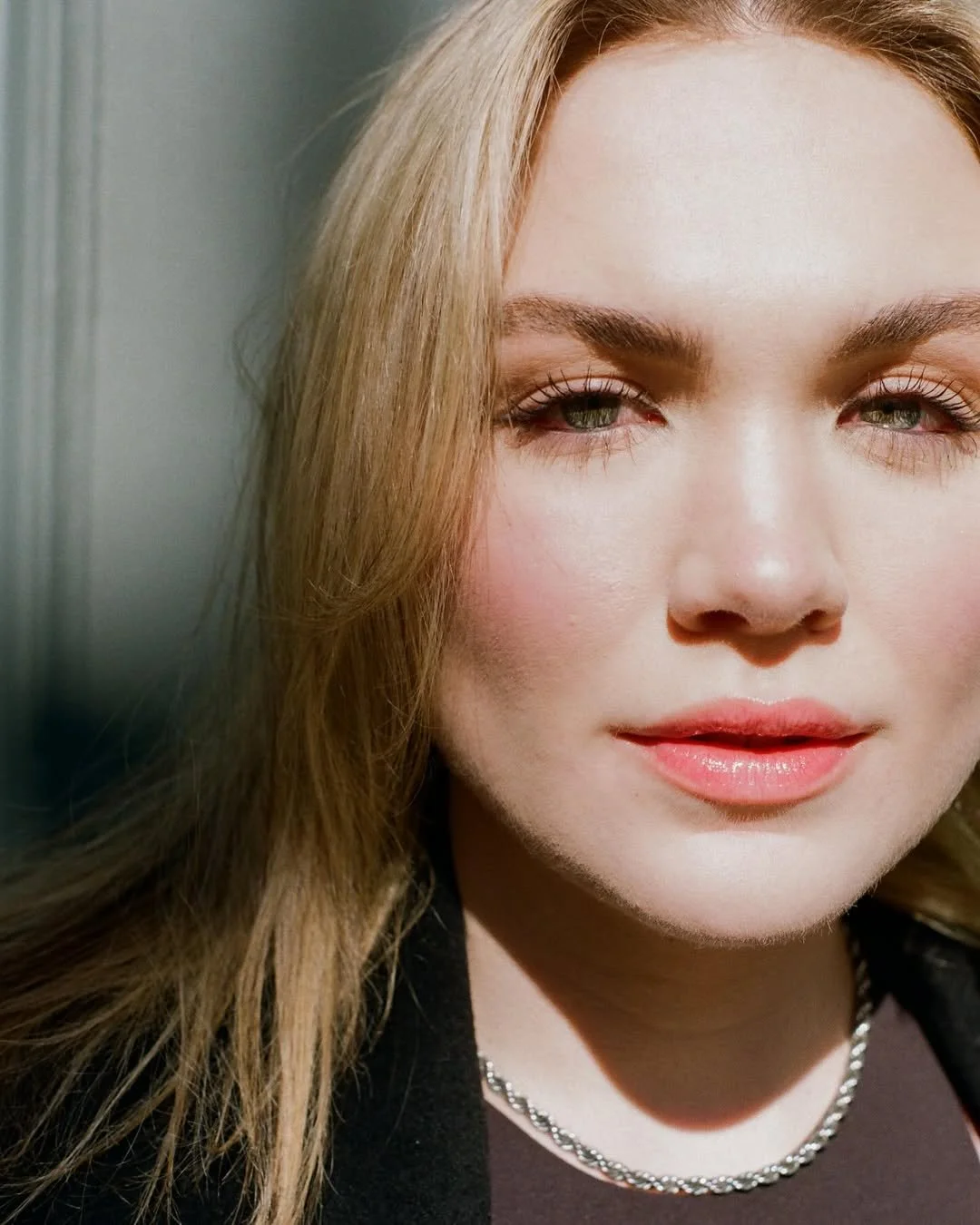 A close-up of a woman with long, light brown hair and fair skin. She has green eyes, well-defined eyebrows, and wears light makeup with pink lipstick. She is wearing a black top and a silver chain necklace.