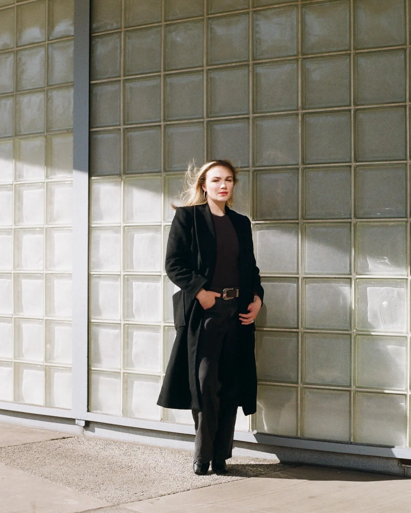 A woman dressed in black, standing in front of a glass block wall on a sidewalk.