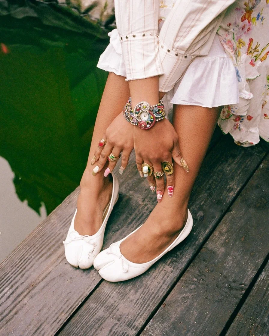 Person wearing white ballet flats and colorful jewelry, sitting on a wooden dock with their hands resting on their knees, near a body of water surrounded by green lily pads.