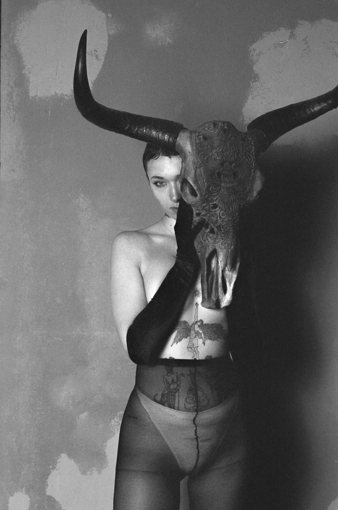 Black and white photo of a woman holding a large animal skull with horns in front of her face. She is wearing long black gloves, a sheer skirt, and has tattoos on her torso and thighs. One eye is visible, peeking from behind the skull.