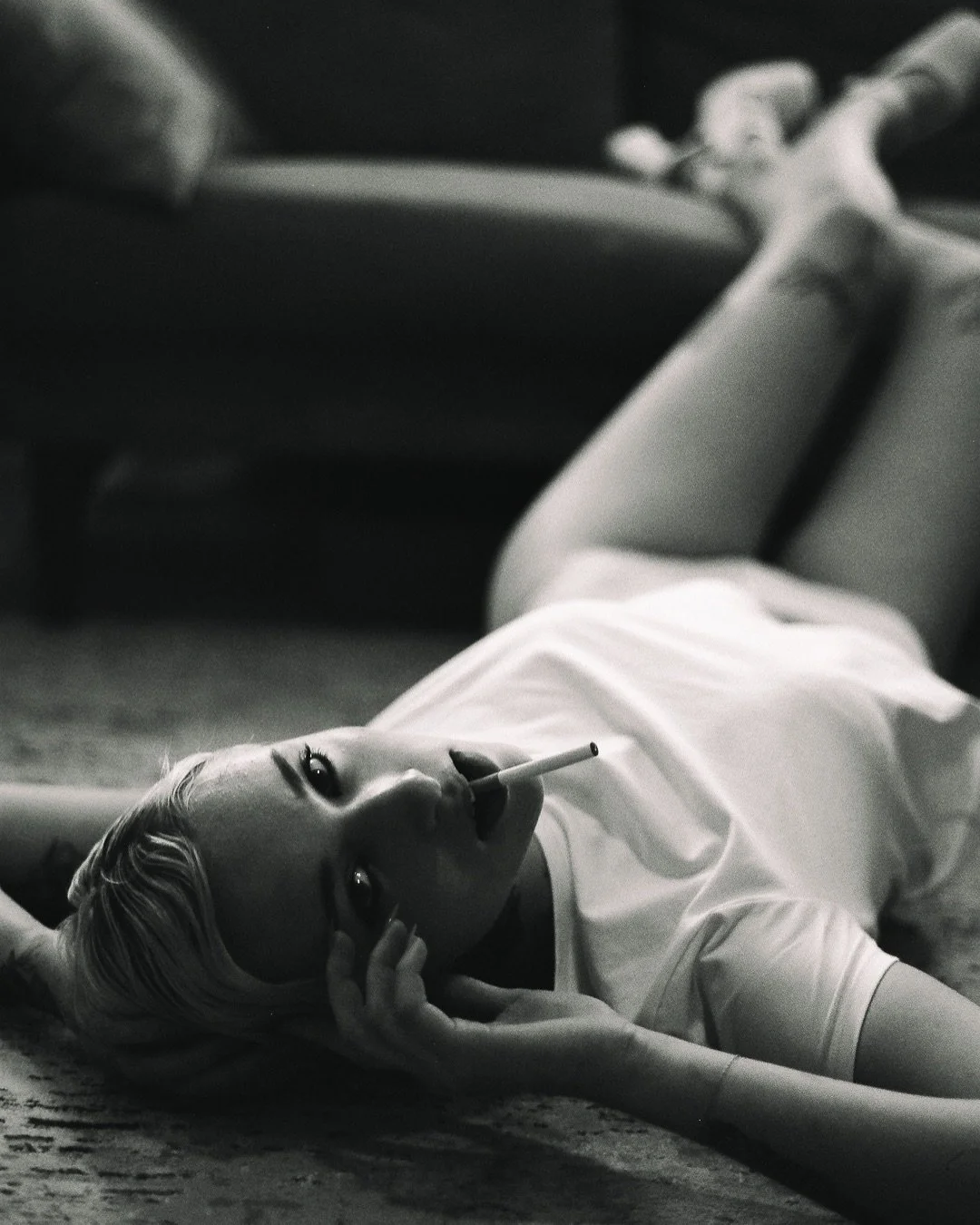 A woman with platinum blonde hair lying on the floor, smoking a cigarette, wearing a white dress, with her head resting on her hand, in a black and white photograph.