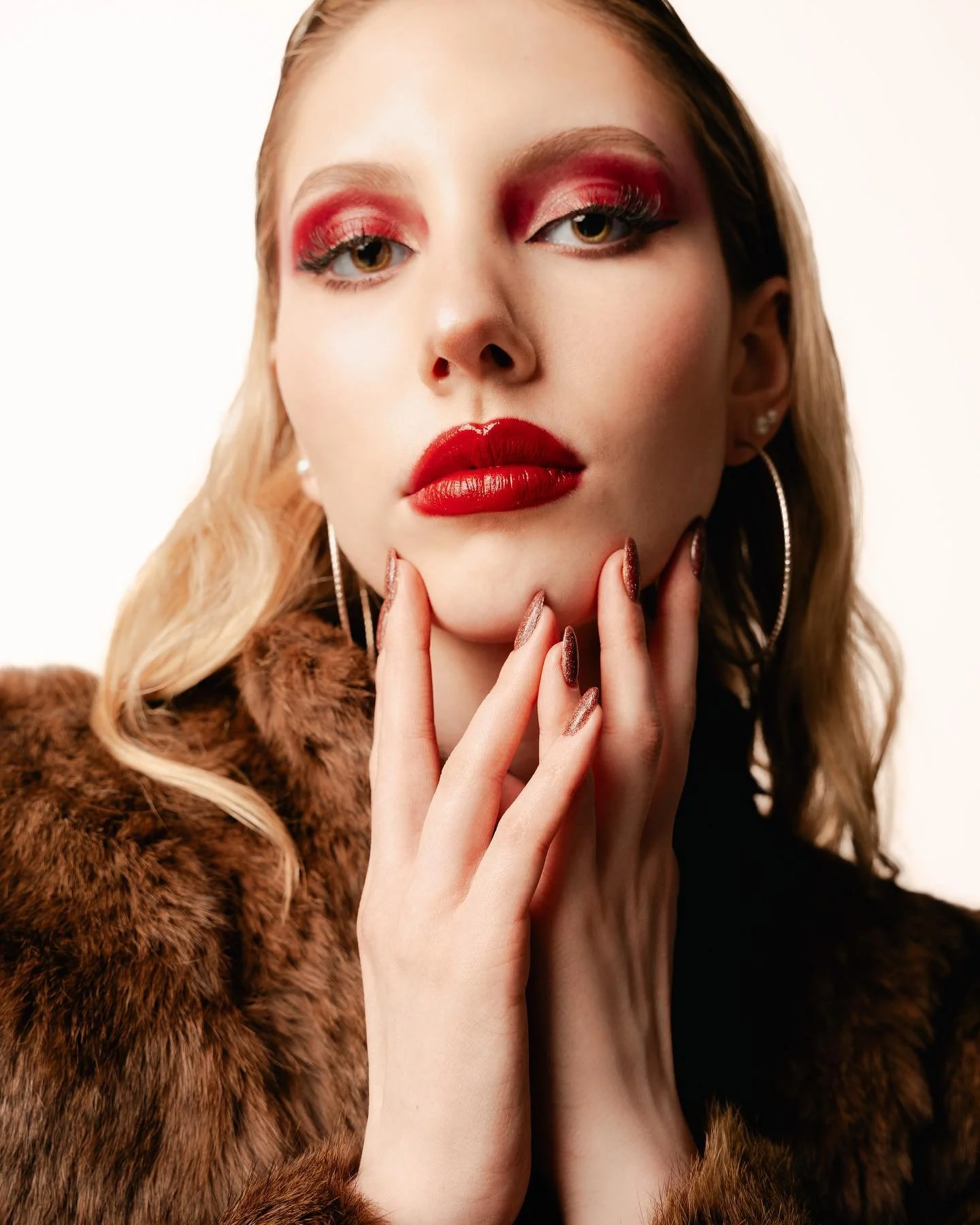 A woman with blonde hair and makeup, wearing a fur coat and large hoop earrings, touches her face with manicured nails.
