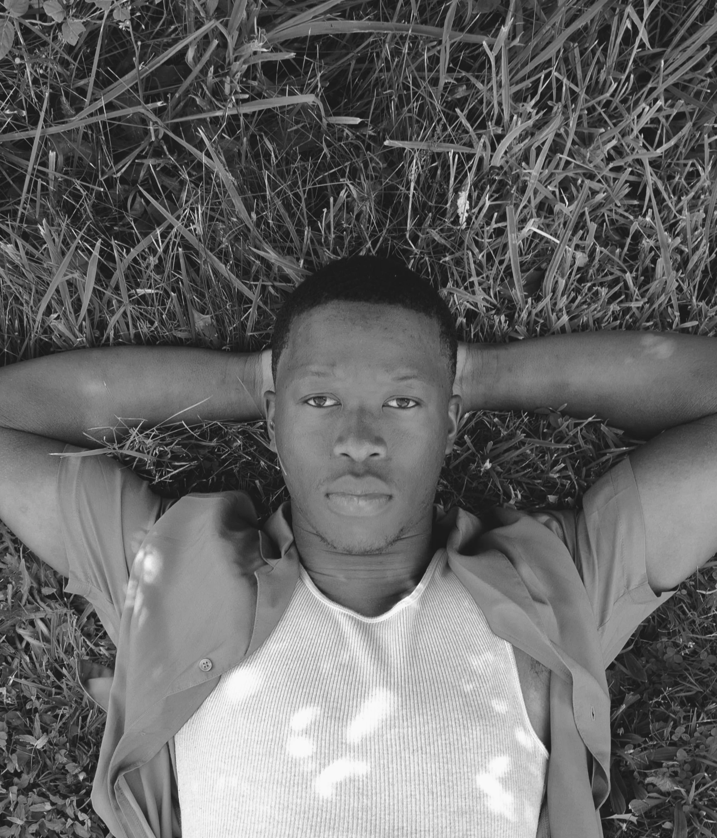 A young man with short hair lying on grass, looking into the camera with his arms stretched above his head and his face centered in the image.