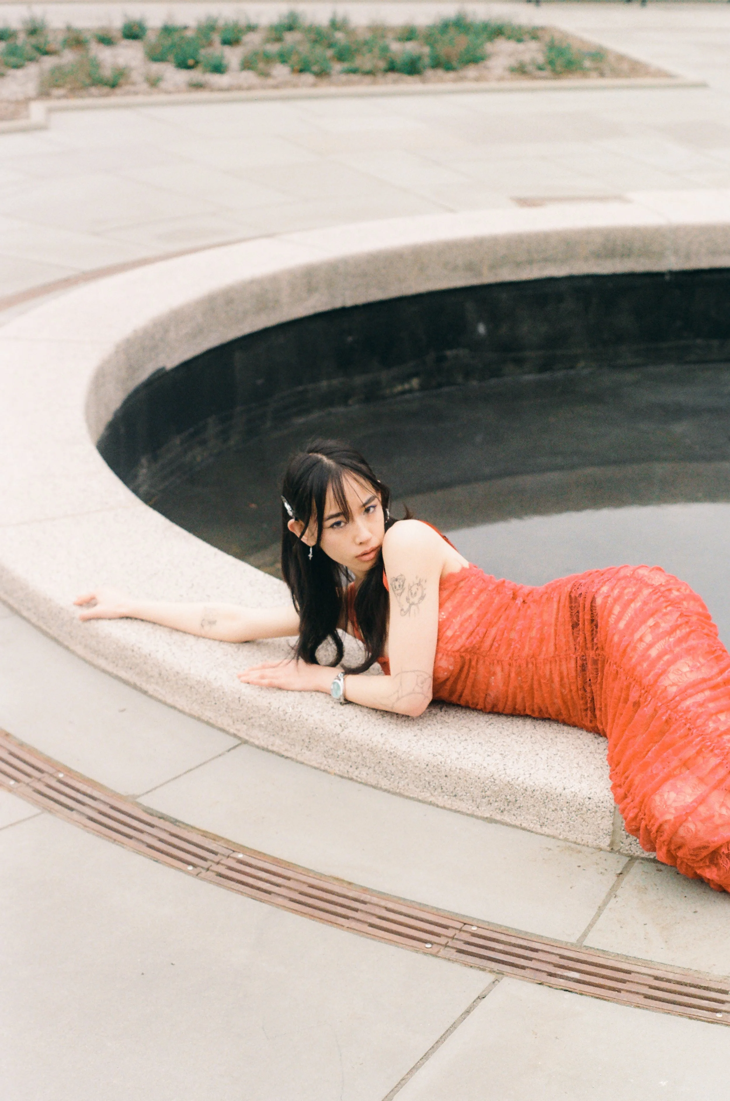 A woman in an orange dress lying on her side on a concrete bench adjacent to a round water feature with dark water, in an outdoor urban setting.