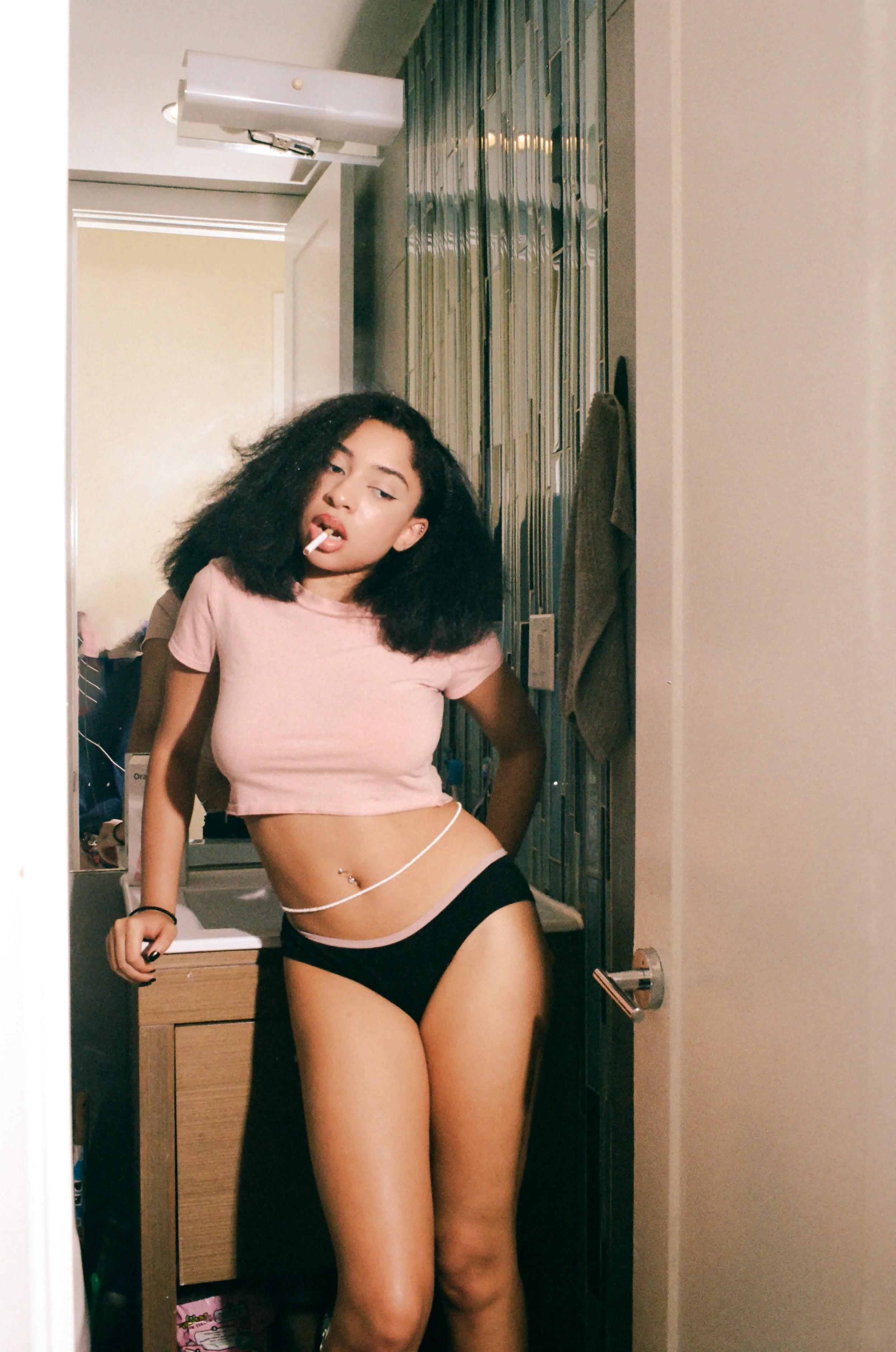 A young woman with curly black hair is standing in a small room, wearing a pink crop top and black shorts, with a cigarette in her mouth. She is leaning against a door frame, with a hand resting on furniture, in a casual pose.