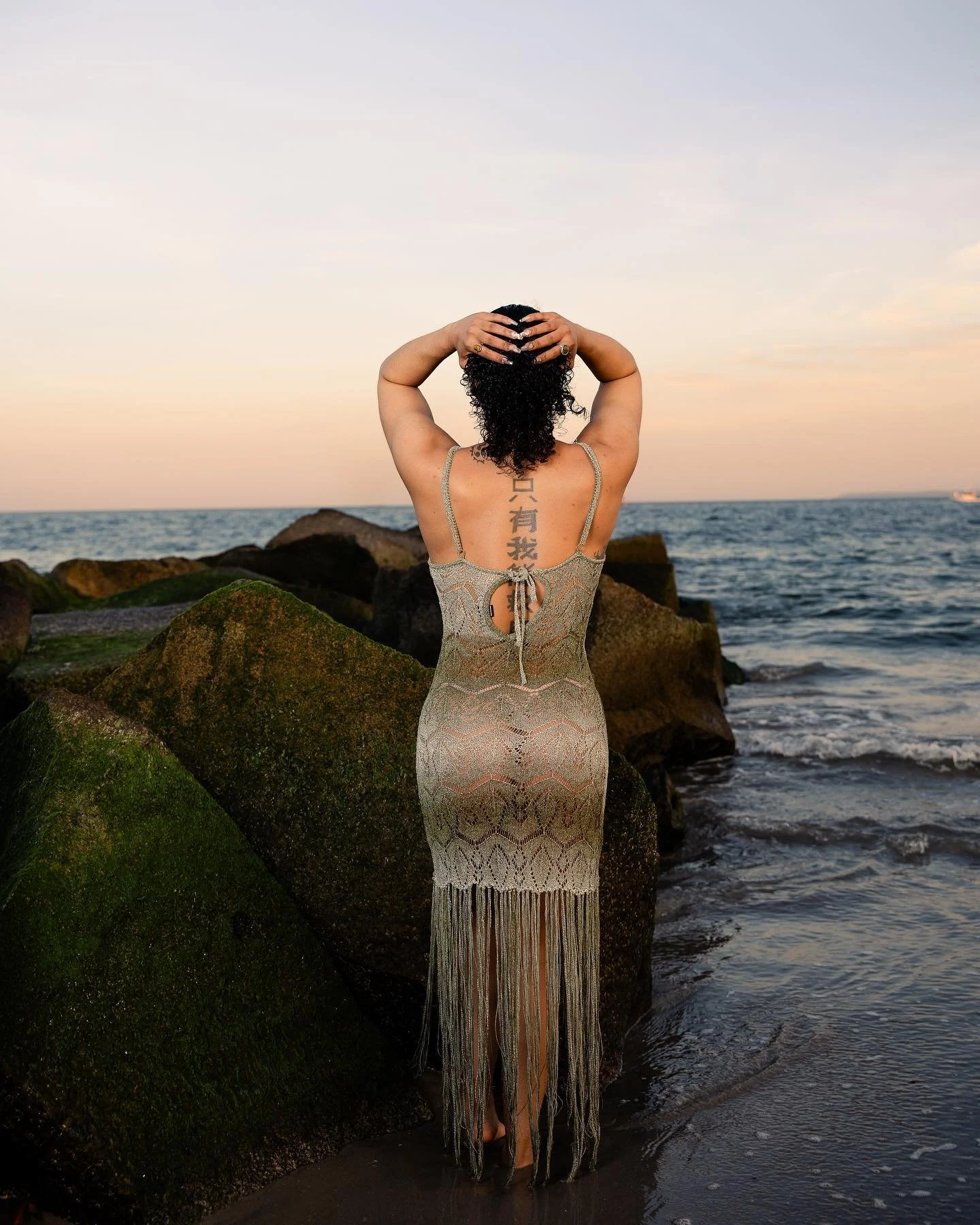 A woman with curly black hair standing on a beach, facing the ocean at sunset, wearing a beige lace dress with fringe details and a tattoo with Chinese characters on her back.