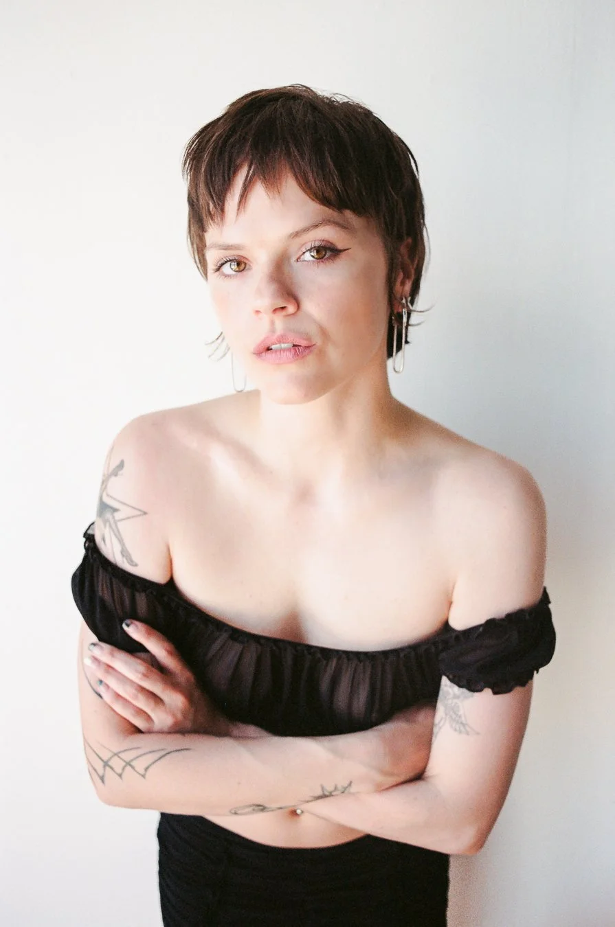A woman with short brown hair, wearing an off-the-shoulder black top, has tattoos on her arms, and is standing against a plain white background.