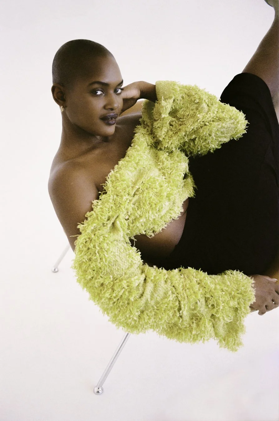 A woman with short hair and dark lipstick sitting on a chair, wearing a yellow fuzzy sweater and black bottoms, against a plain white background.