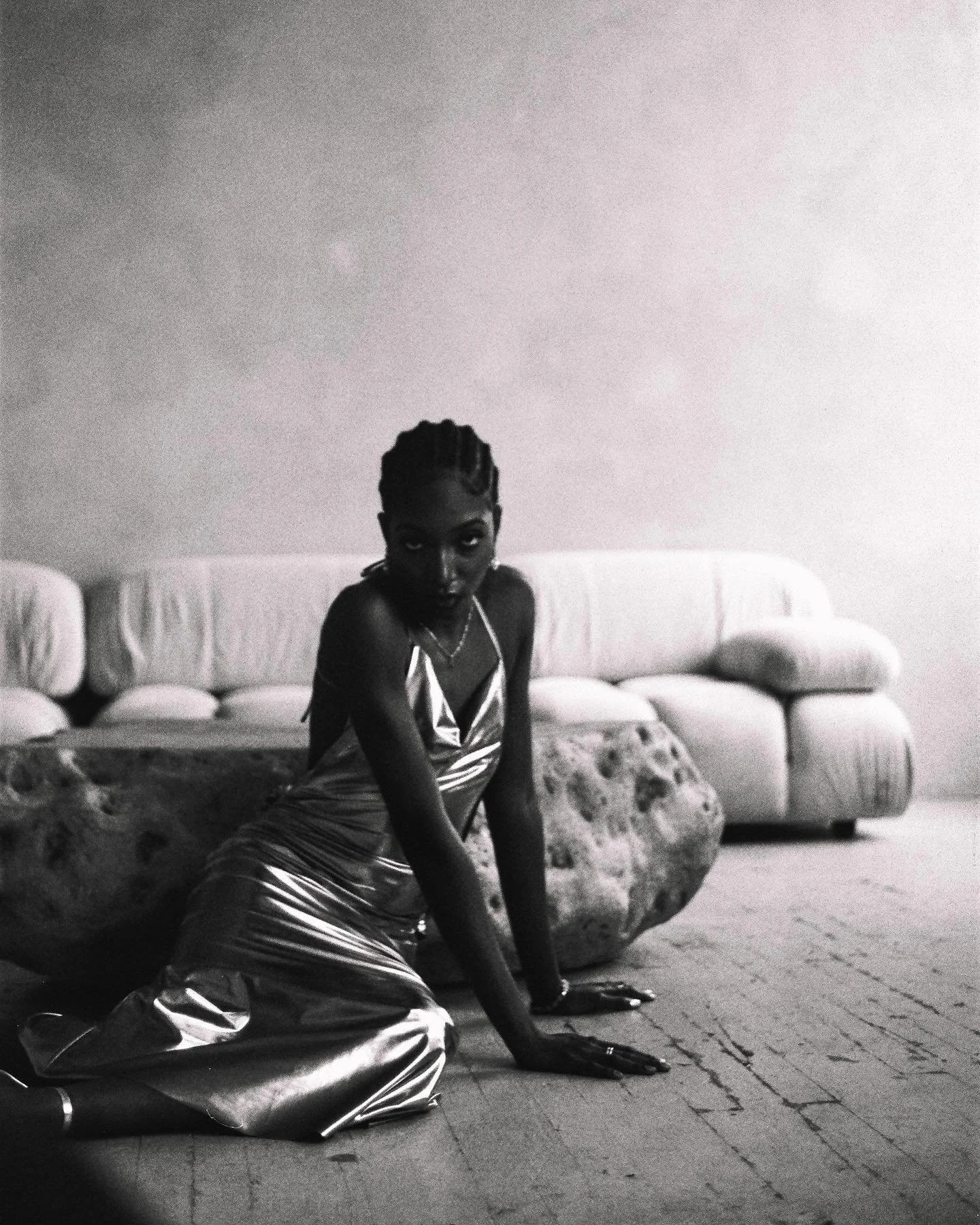 A woman with braided hair and dark skin is sitting on the floor next to a large, textured rock. She is wearing a shiny, metallic dress with thin straps. Behind her, there is a light-colored, curved sofa with rounded armrests and several cushions. The