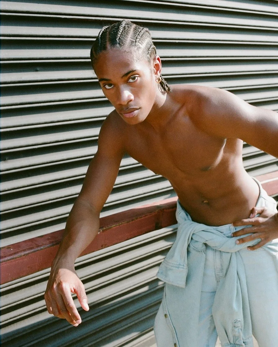 A young man with braided hair leaning against a metallic wall, shirtless with a light-colored jacket tied around his waist.