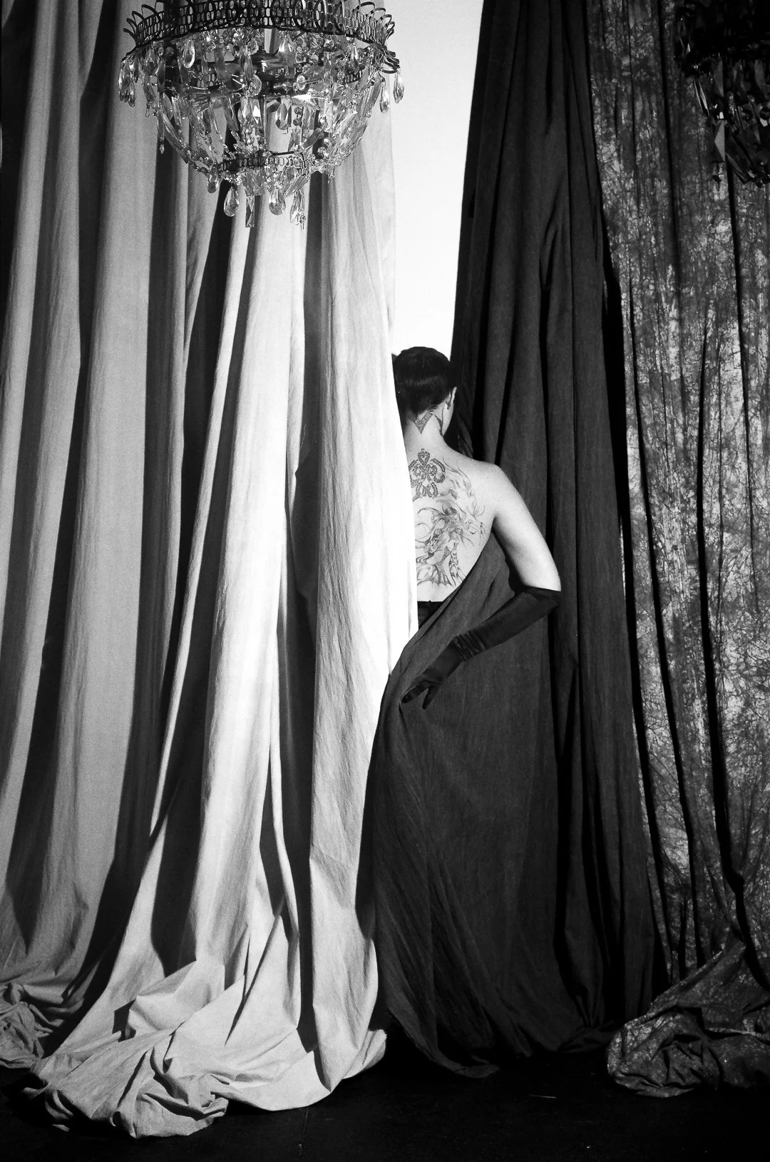 A woman with a large tattoo on her back stands partially behind curtains, facing away, in a room with a chandelier and dark curtains.