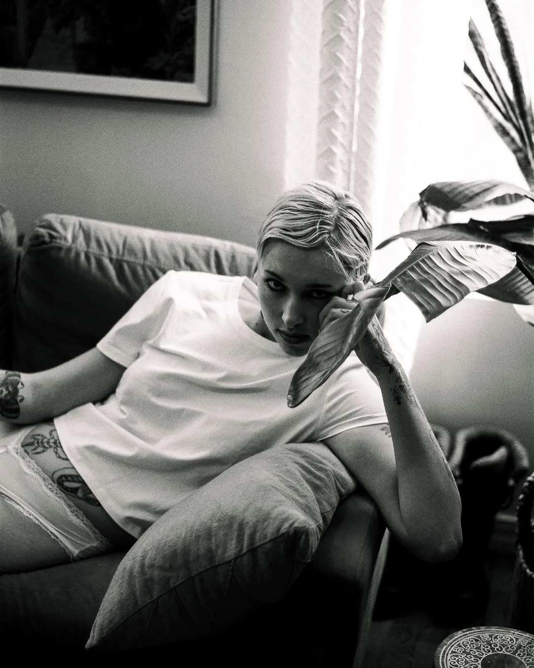 A black and white photo of a woman lounging on a couch, holding a large leaf and looking at the camera with a serious expression.