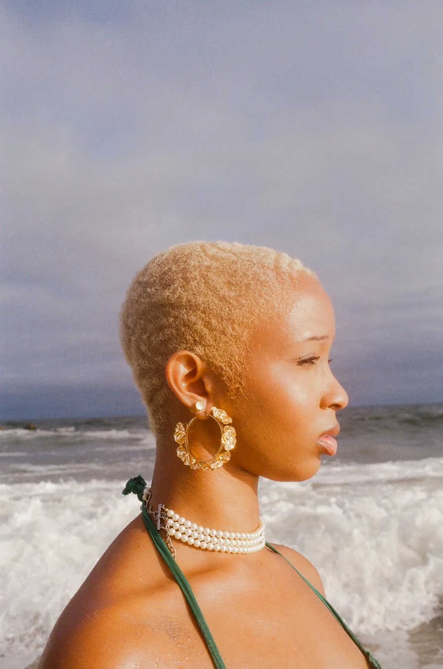 Side profile of a woman with short blonde hair, wearing pearl earrings, a pearl necklace, and a green bikini strap, standing on the beach with the ocean and cloudy sky in the background.