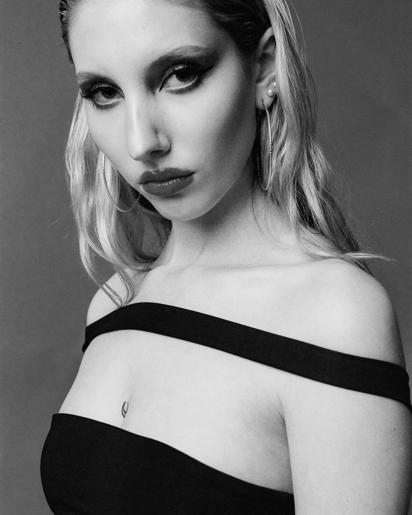 A black and white portrait of a woman with light hair, wearing hoop earrings and a strapless top, with intense eye makeup.