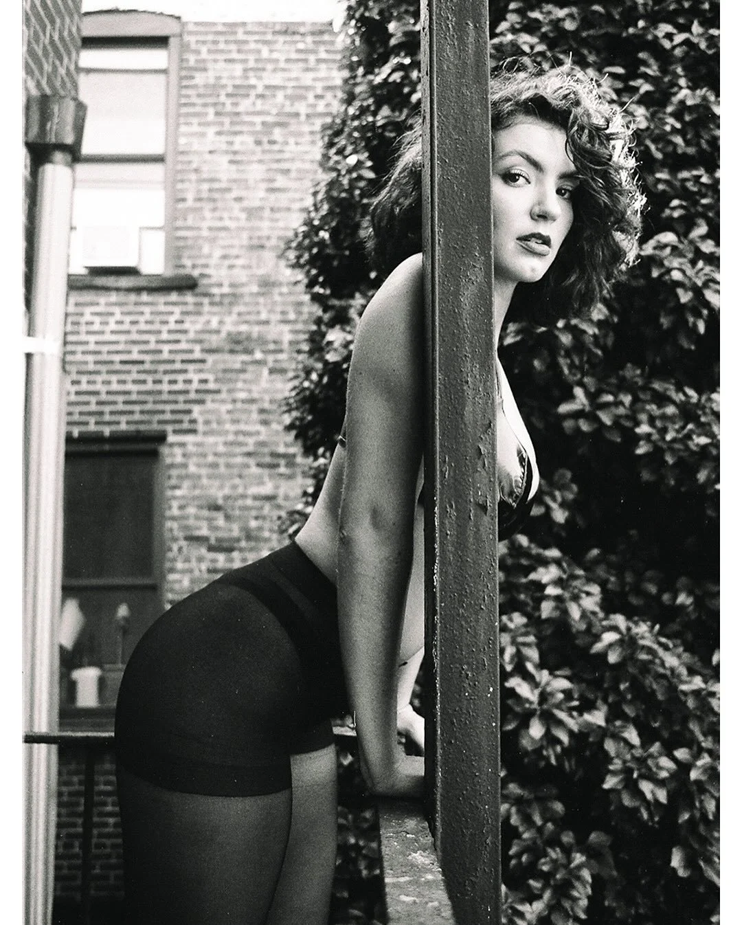 A young woman with curly hair leaning against a pole outdoors, with a brick building and leafy bushes in the background, captured in black-and-white.