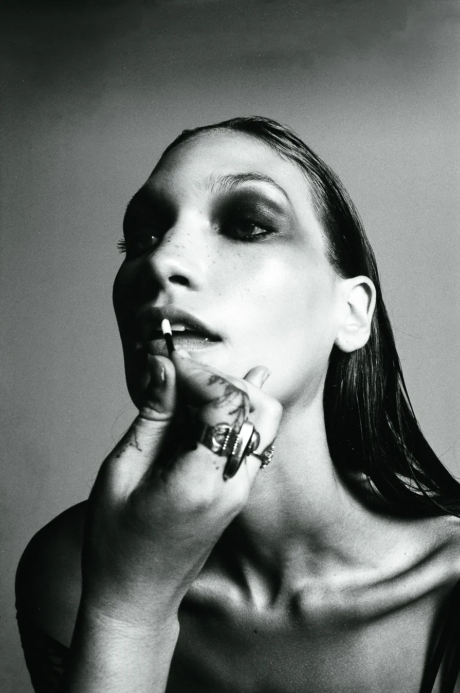 Black and white photo of a woman applying lipstick, with dark eye makeup, wet hair, and wearing rings on her hand.