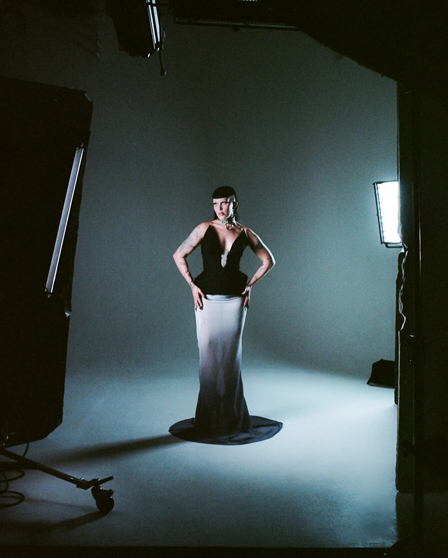 A woman standing on a photo studio backdrop, posing for a photoshoot, wearing a black top and long white skirt, with studio lights set up around her.