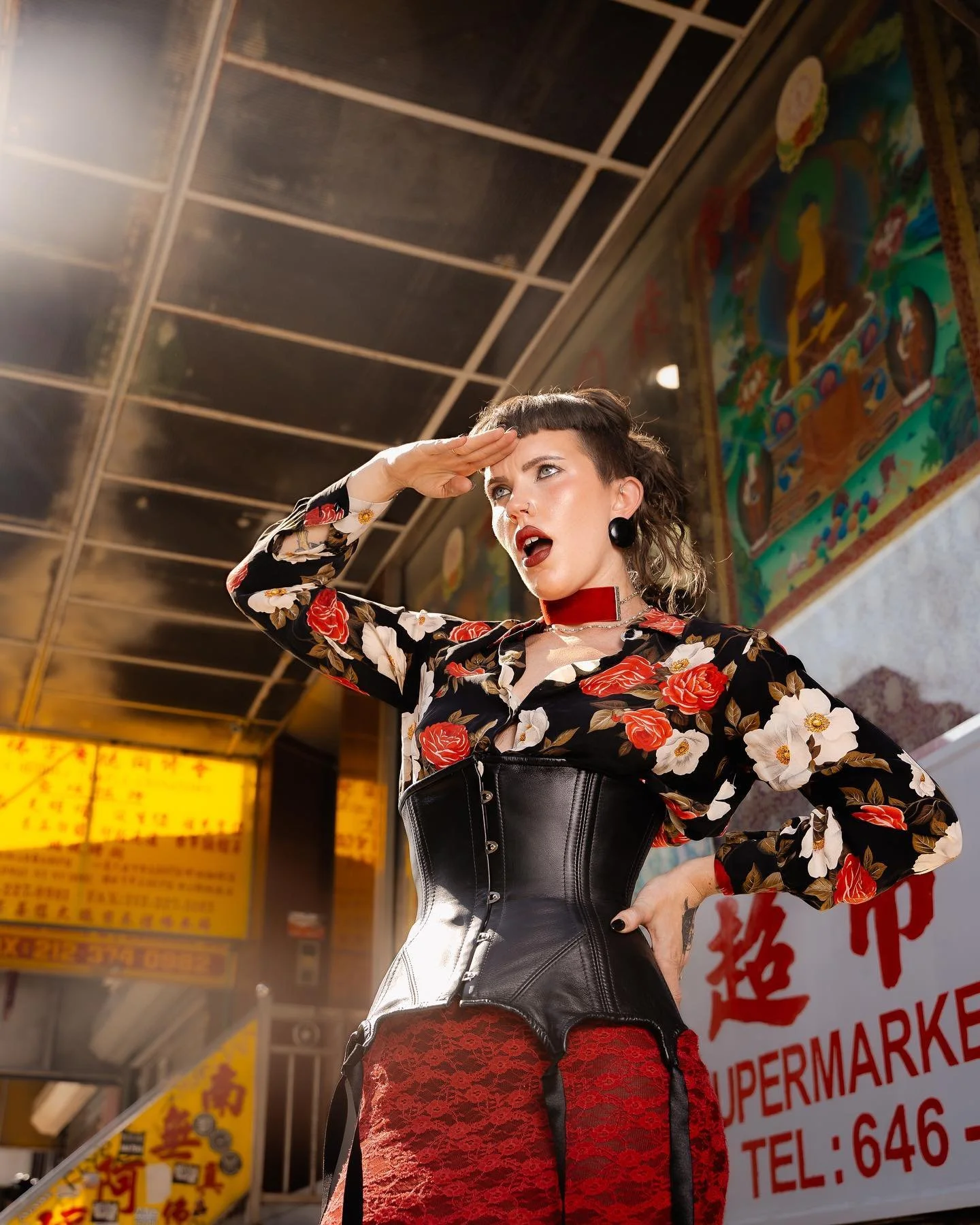 A woman with short hair, wearing a floral blouse, red lace skirt, black corset, and accessories, standing in front of a storefront with Asian script signs and colorful murals, saluting with her right hand.