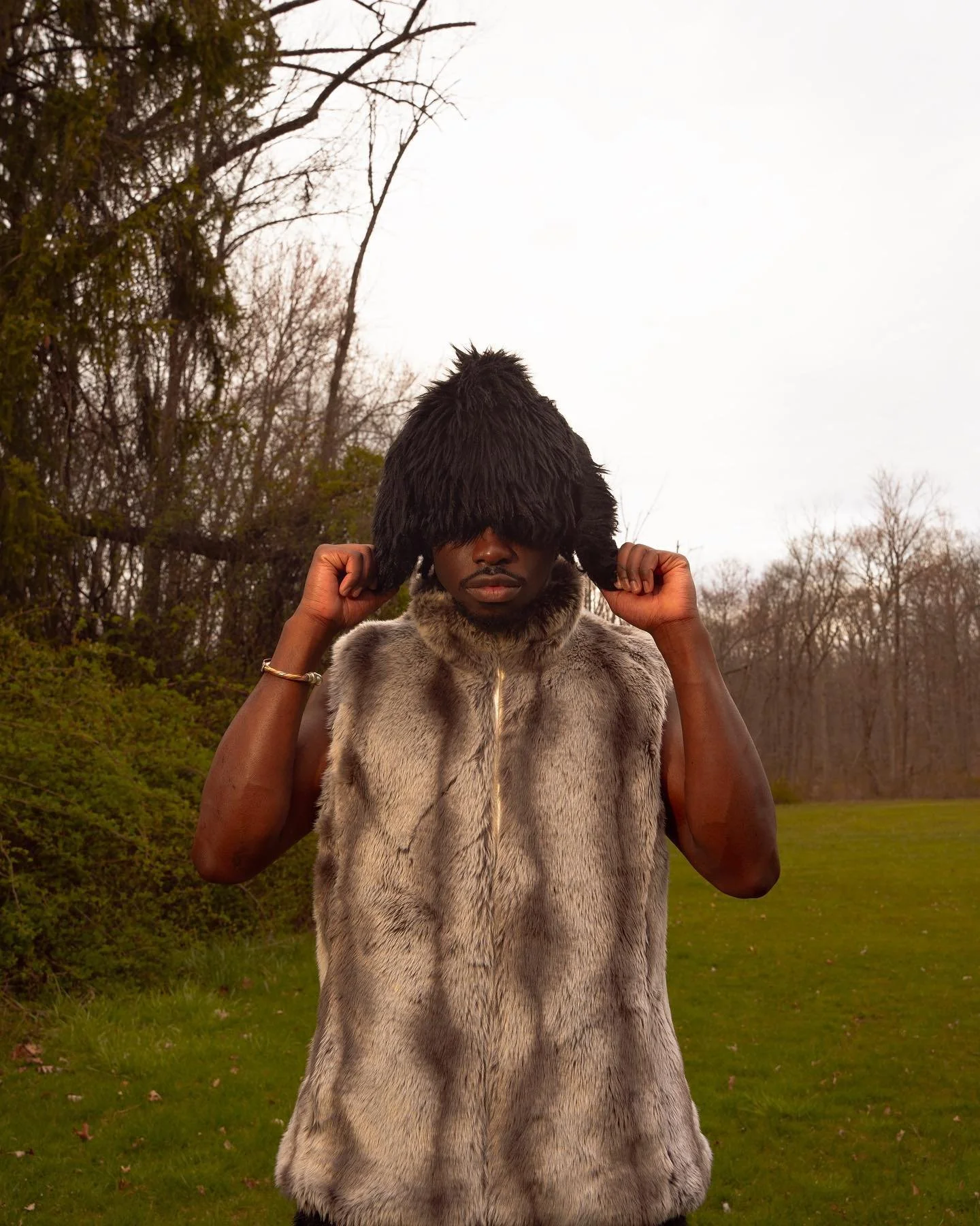 A person standing outdoors wearing a fur vest and covering their eyes with large, furry, black ear-like accessories, against a background of trees and grass.